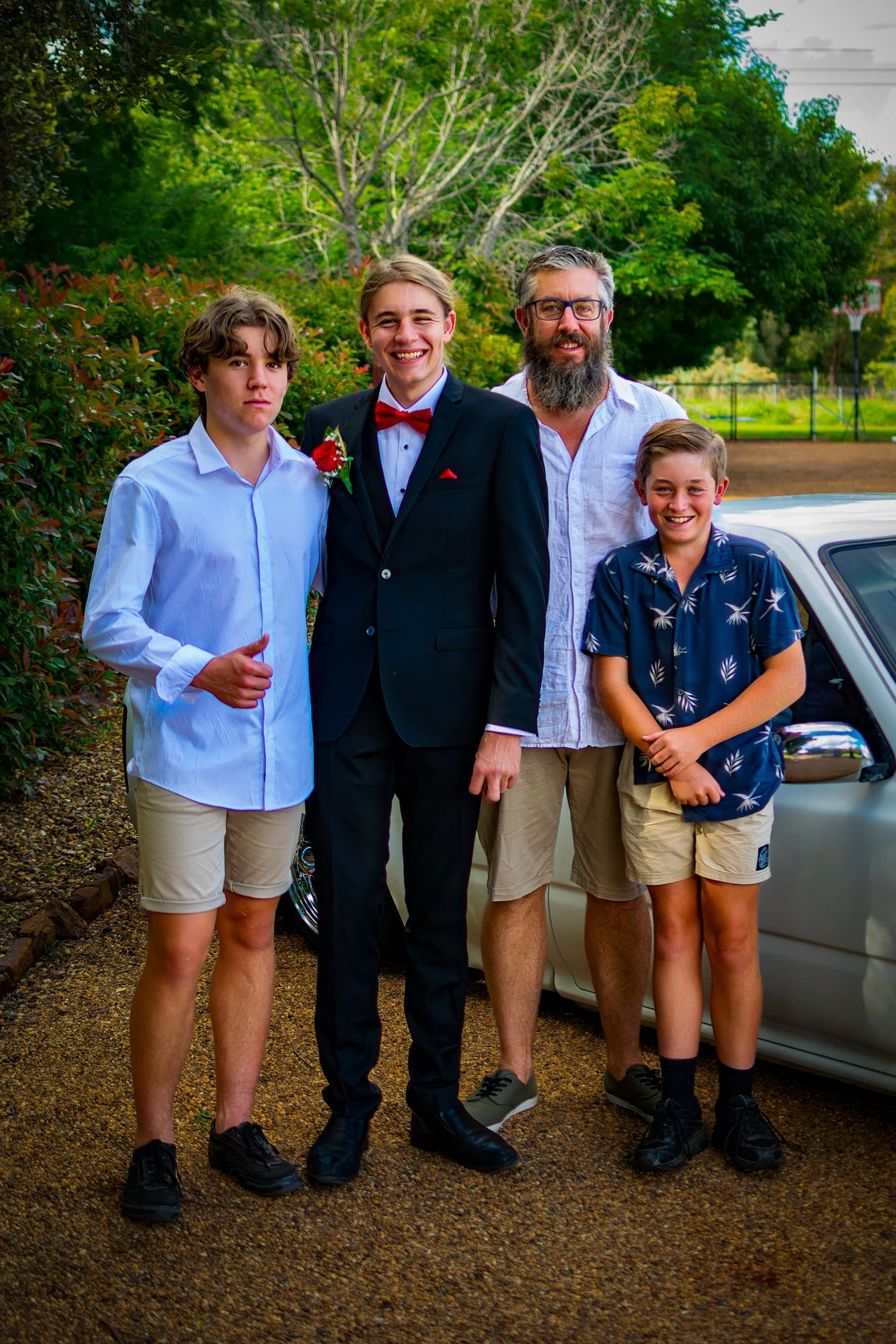 Group Of Four Posing Outdoors By A Car; One Person In A Suit, Others Casual.