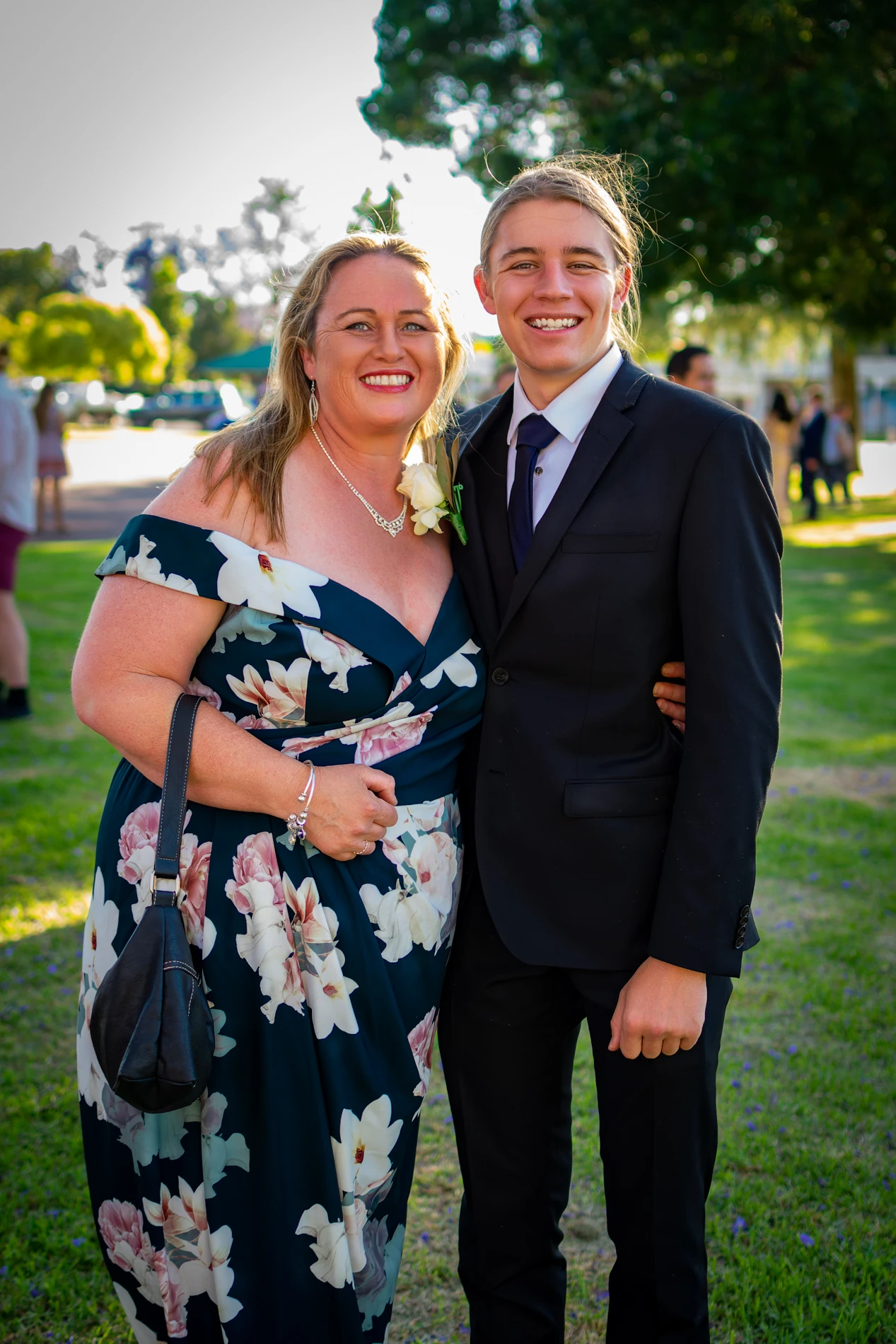 Adult In Floral Dress And Young Person In Suit Smiling Outdoors At A Formal Event.