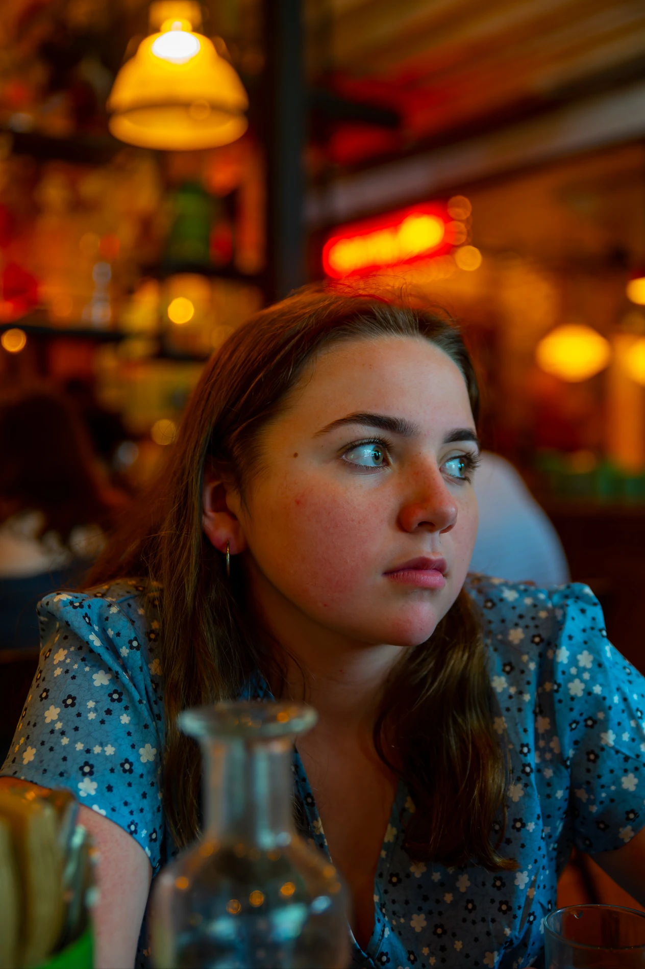 Young Woman In Blue Floral Dress Looking Right, Dim Cafe With Warm Blurred Lighting.