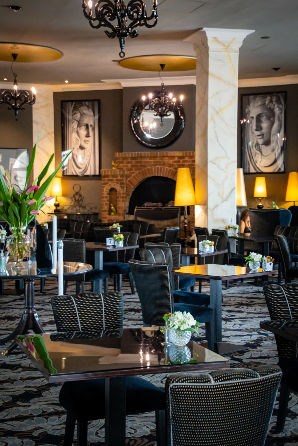 Stylish Indoor Dining Area With Dark Furniture, Floral Decor, Wall Art, Chandeliers, Fireplace.