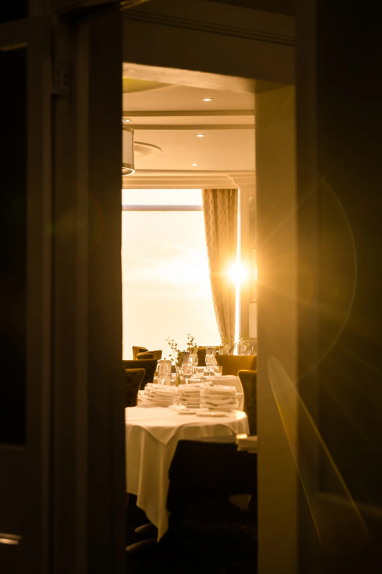 Warmly Lit Dining Room With Set Tables And Sunlight Through The Window, Viewed Through Doorway.