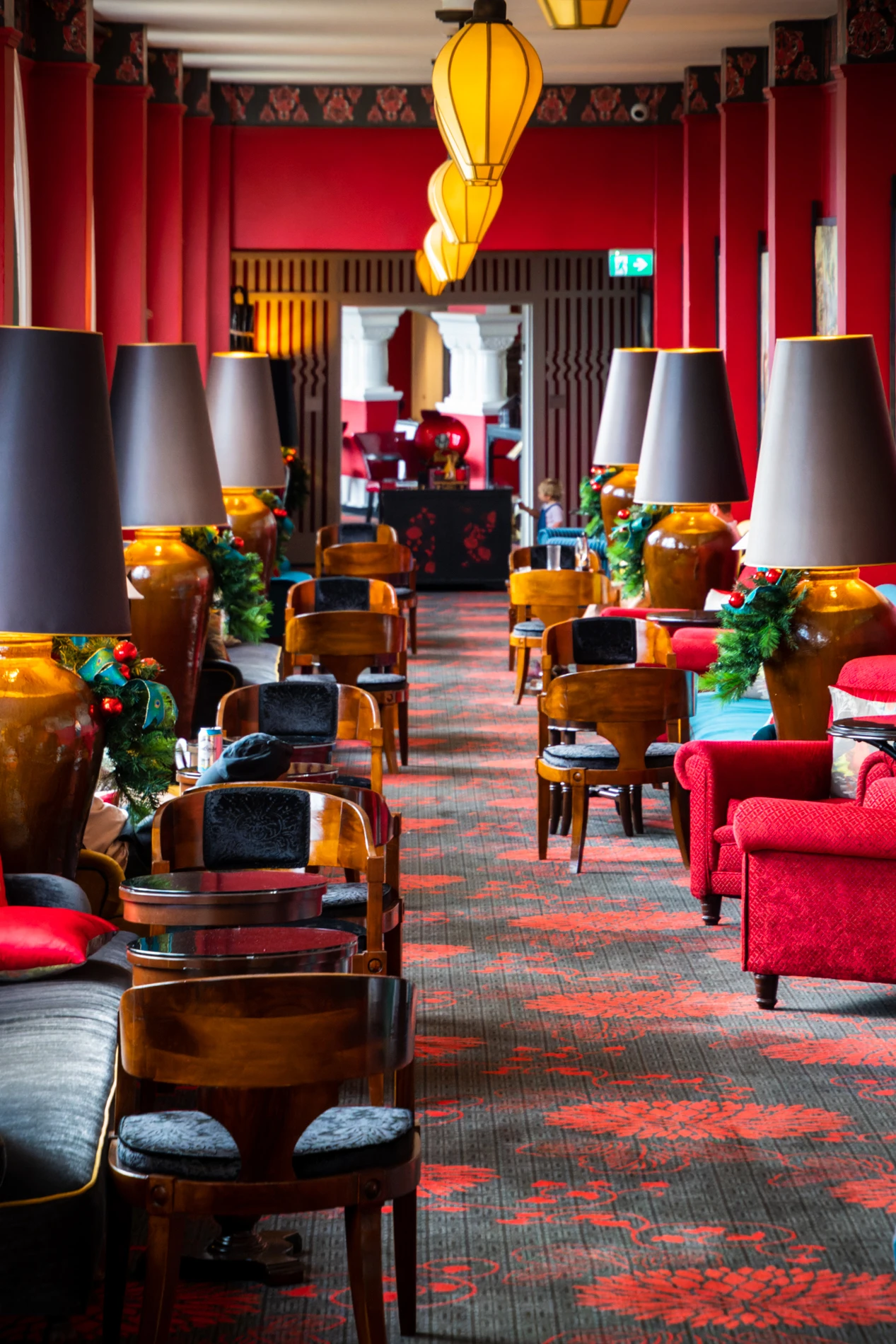 Vibrant Lounge With Red Walls, Floral Carpet, Wooden Furniture, Velvet Seating, And Decorative Lamps.