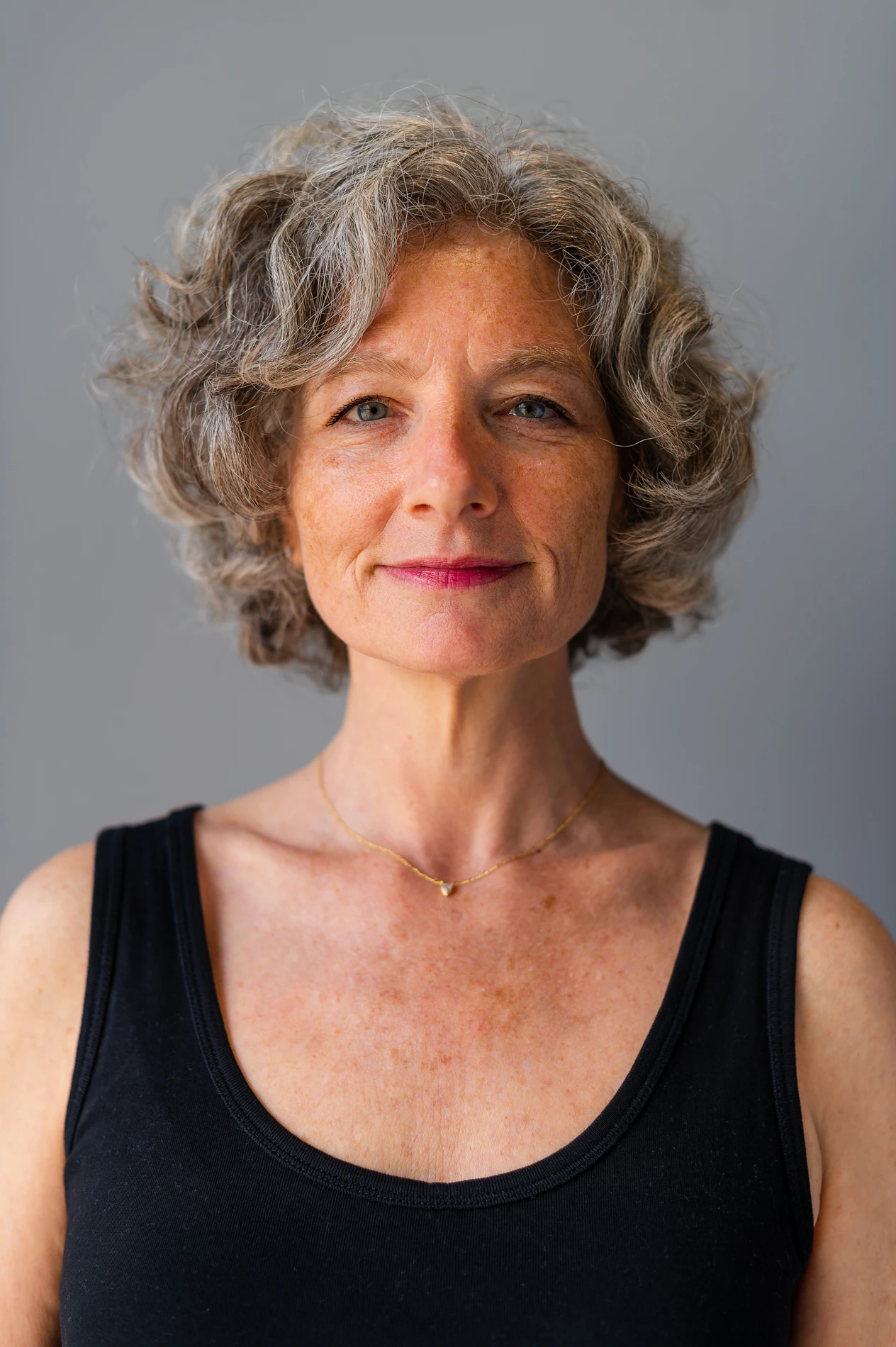 Woman With Curly Grey Hair, Black Top, Gold Necklace Against Plain Background.