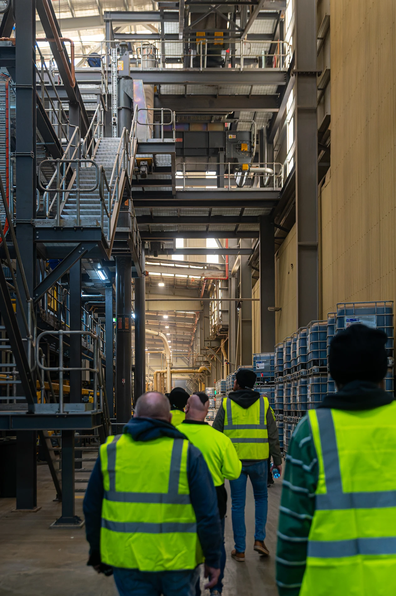 Individuals In High-Vis Vests Walk Through An Industrial Warehouse With Storage Racks.