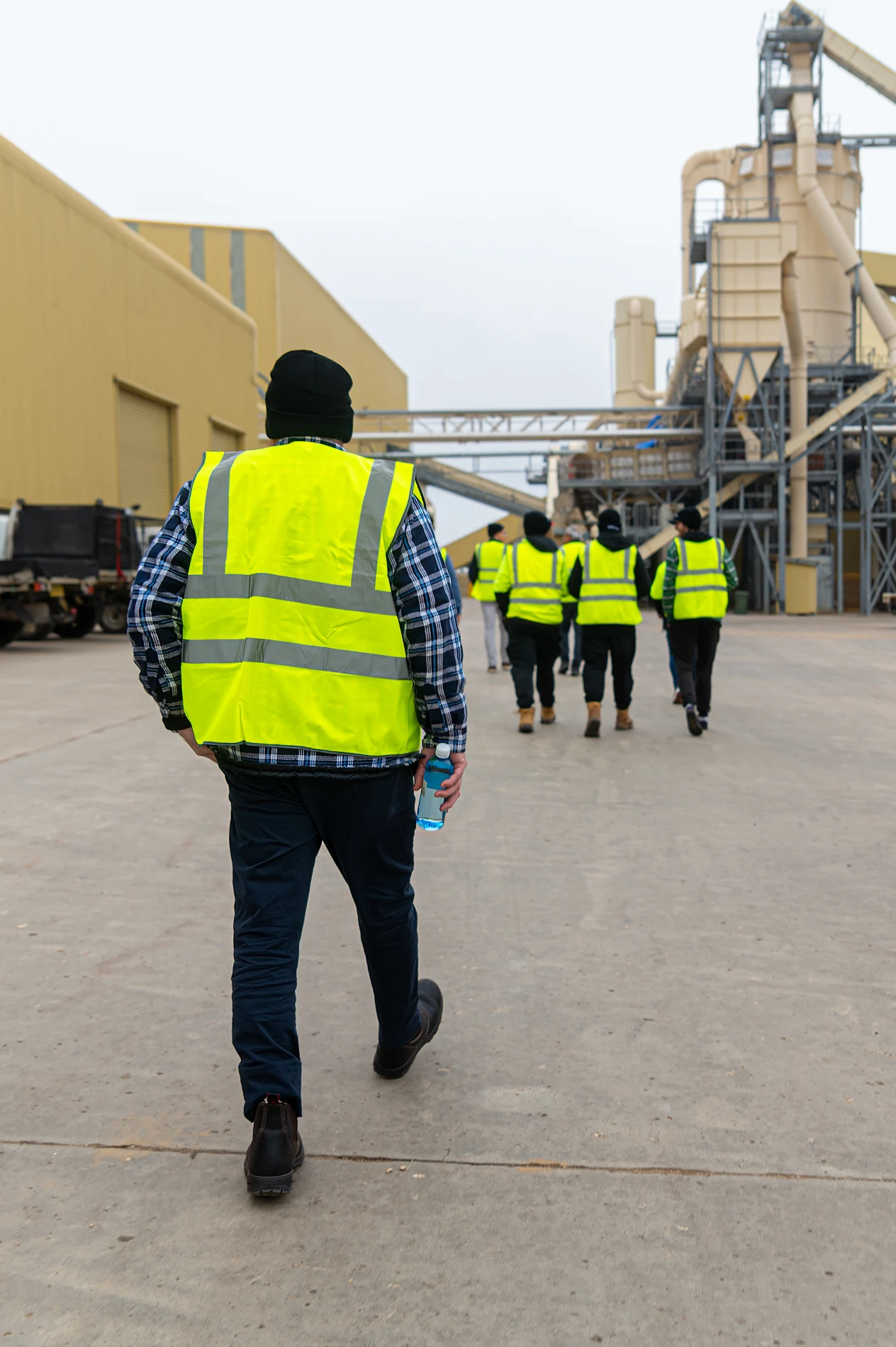 People In High-Visibility Vests Walking In An Industrial Area With Machinery And Buildings.