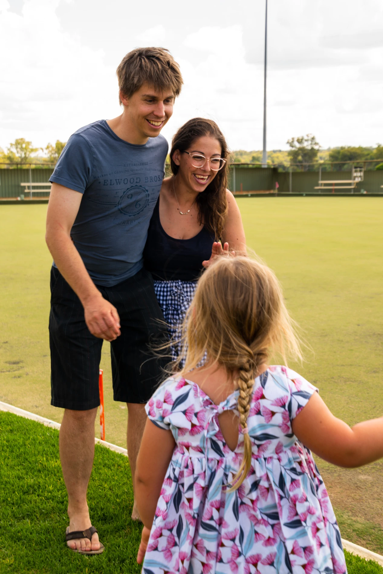 Couple Smiling With A Young Girl In A Floral Dress On A Sunny Lawn Bowling Green.