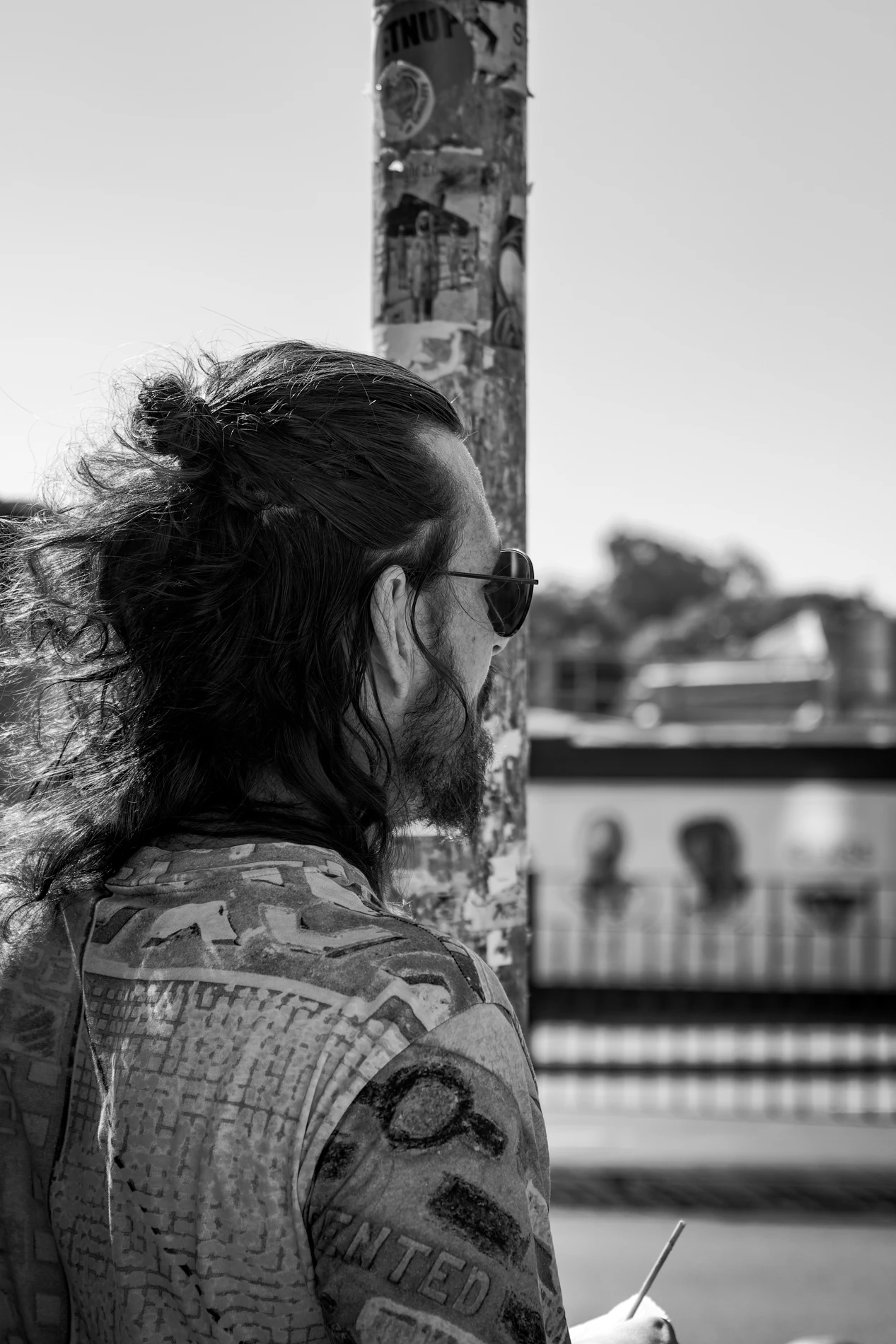 Person With Long Hair, Sunglasses, Patterned Shirt Beside A Poster-Covered Pole In Urban Setting.