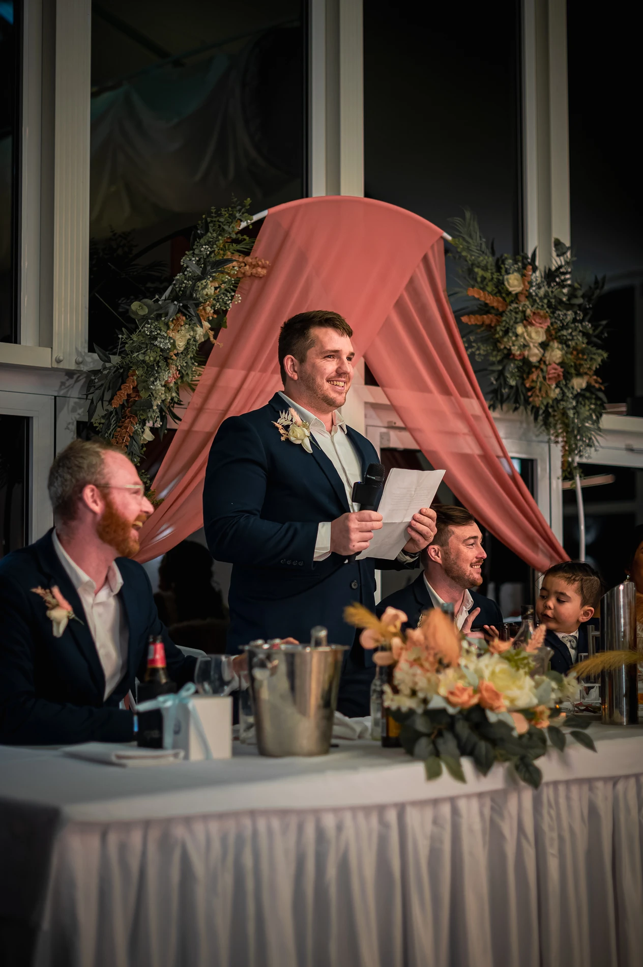 Man In Formal Attire Giving Speech Indoors, Holding Microphone And Paper, By Flower Arch.