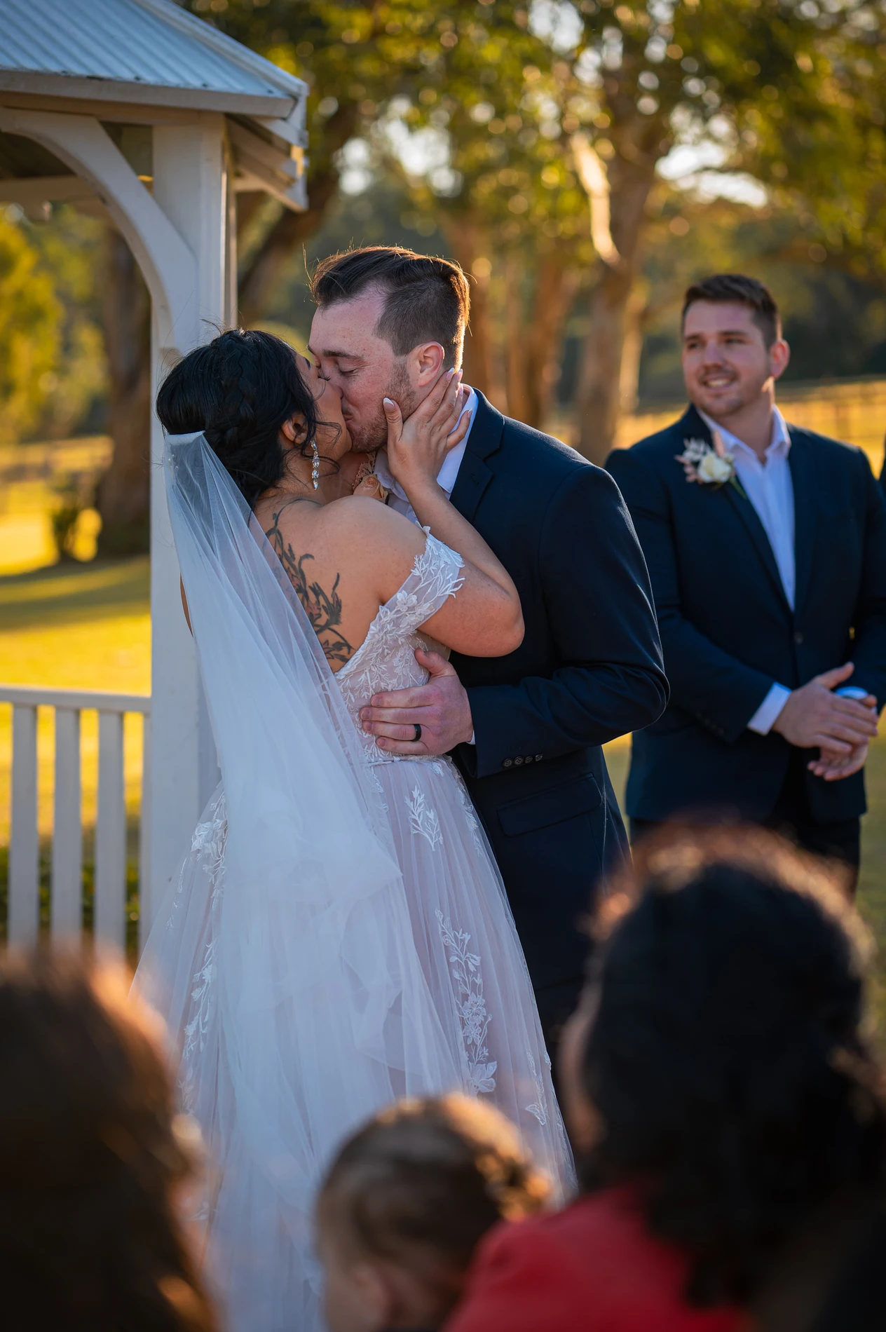 Bride And Groom Kiss At Outdoor Wedding Ceremony With Groomsman Smiling Nearby.
