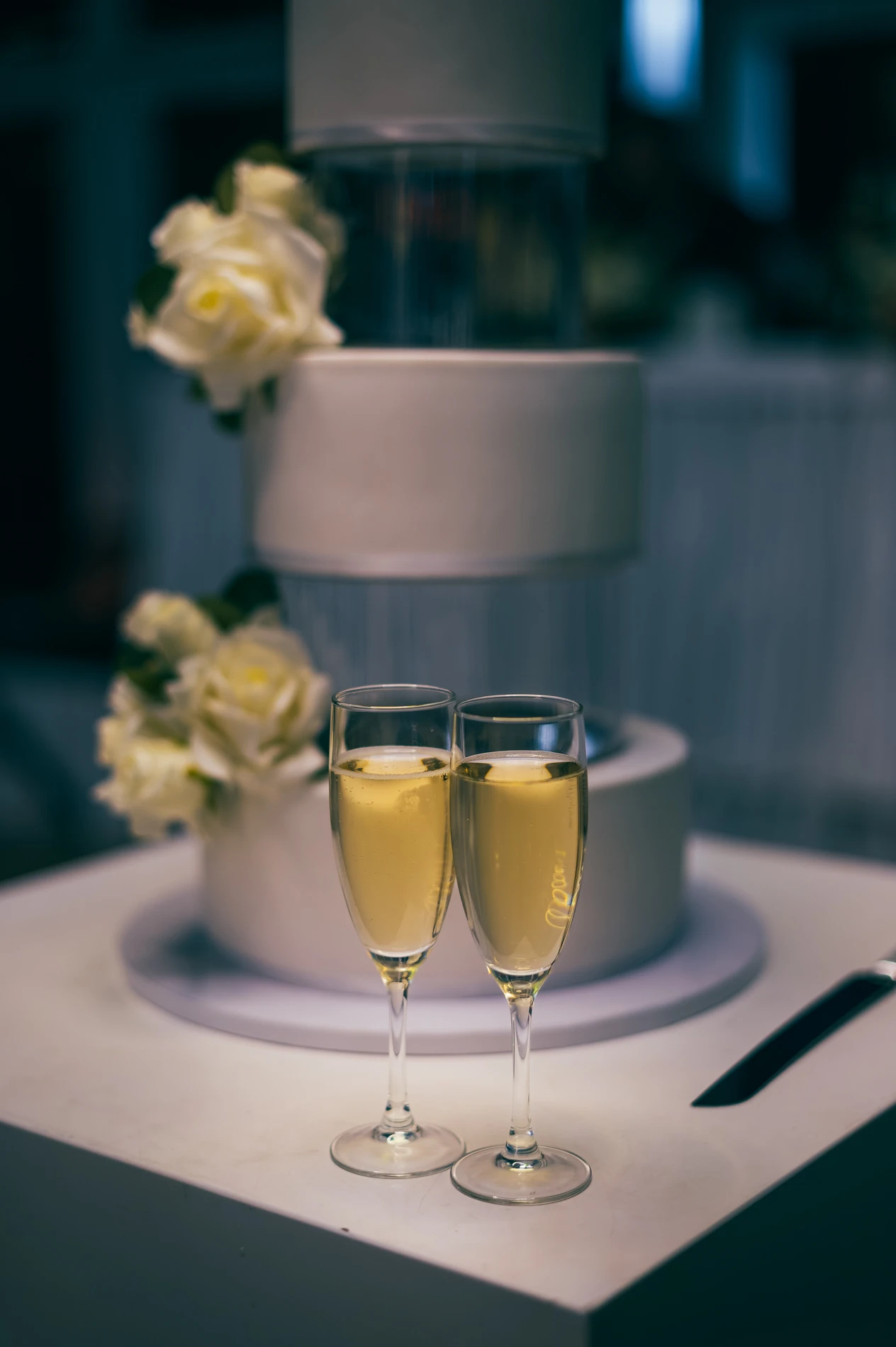Champagne Flutes In Front Of A Tiered White Cake With Flowers; Knife On Table Nearby.