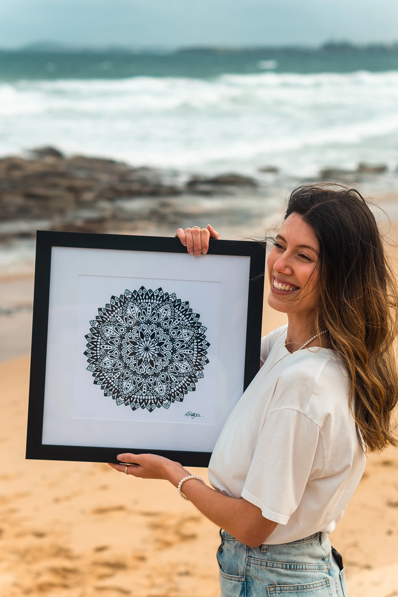 Person On Beach Holding Framed Mandala Artwork; Ocean And Rocks In Background.