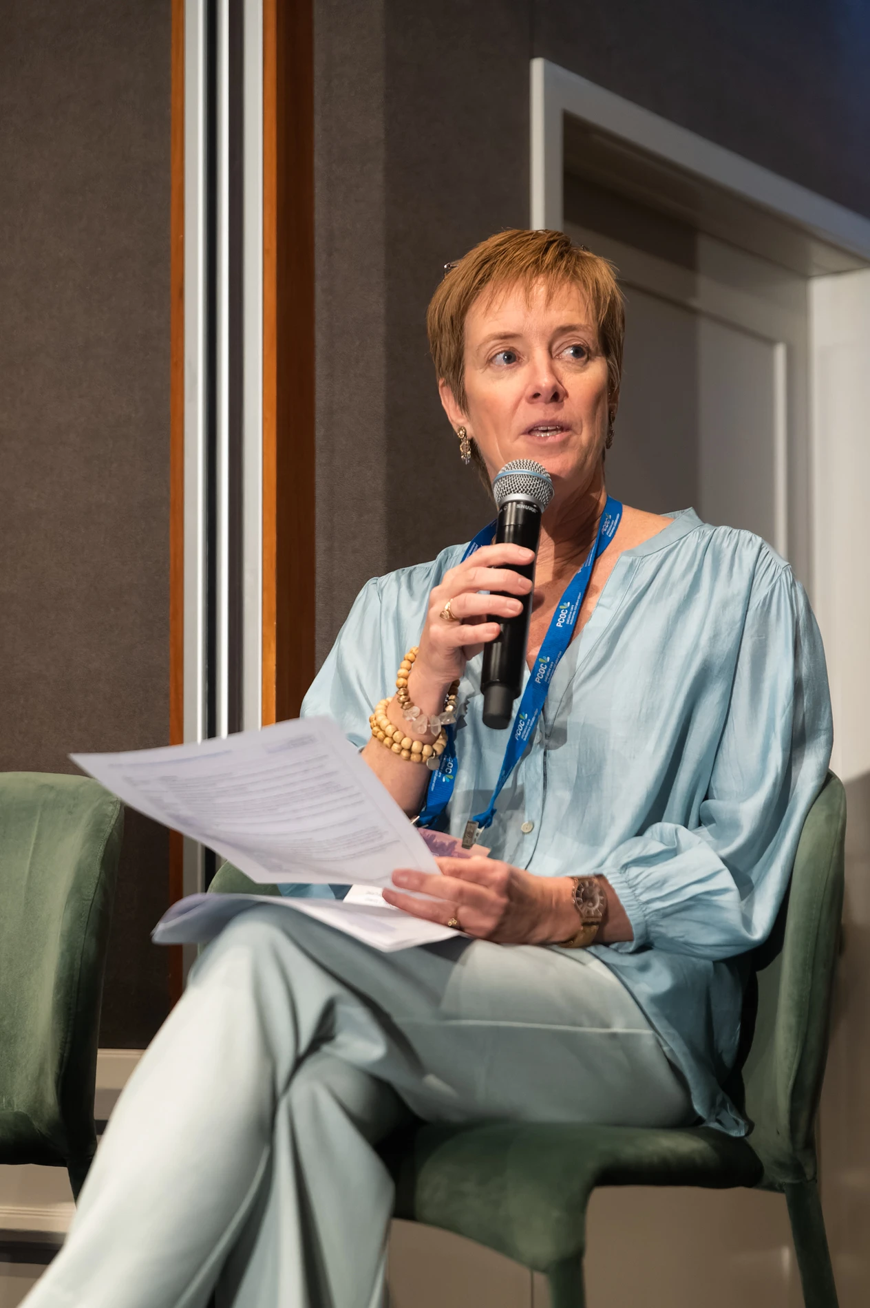 Person Speaking Into A Microphone, Holding Paper, Light Blue Blouse, Lanyard, Indoor Setting.