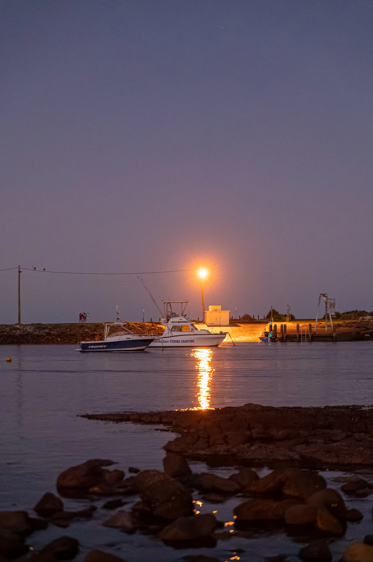 Boats Moored At Dusk, Streetlight Illuminating Pier, Rocky Shore In Foreground.