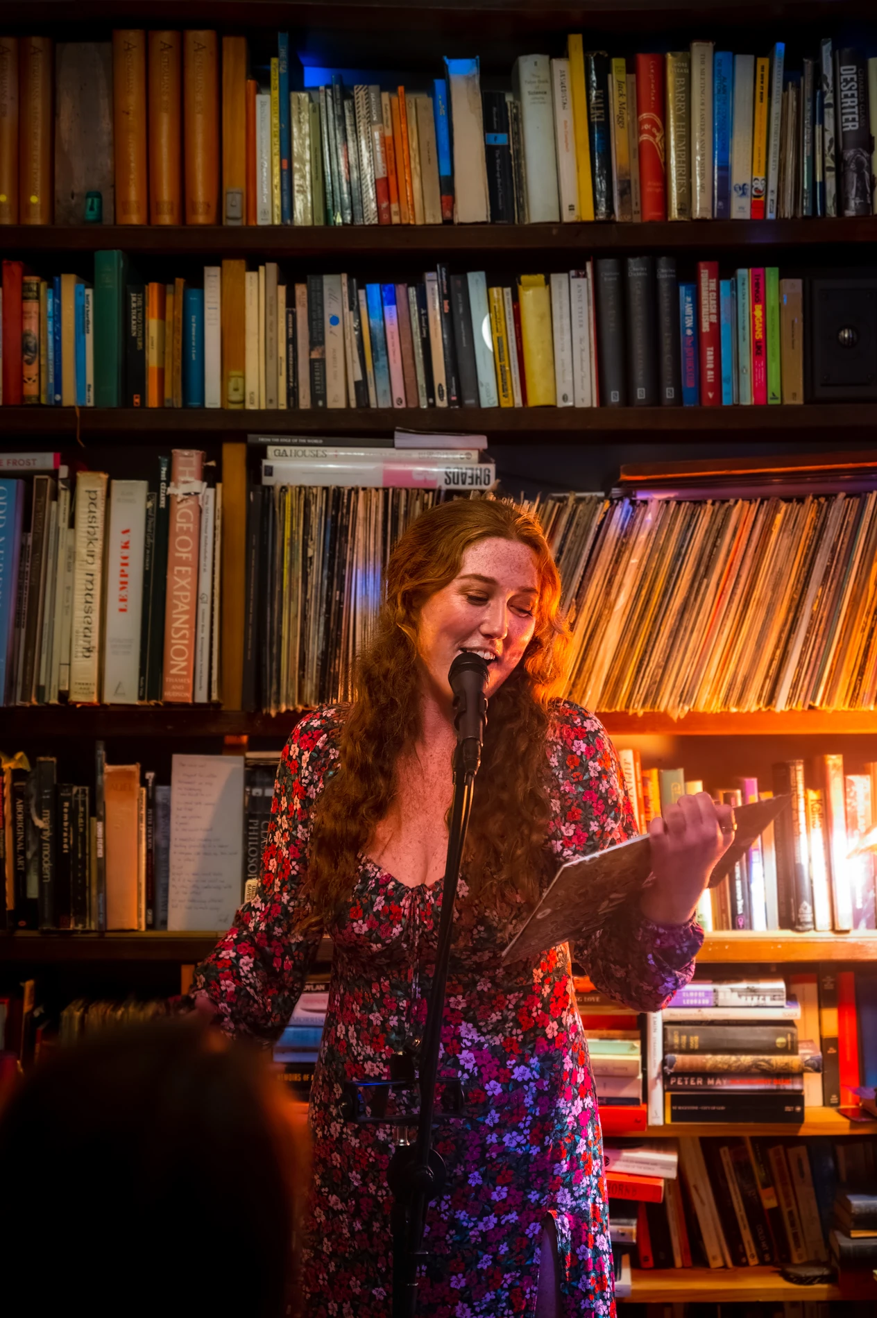 Woman Reads From A Book Into A Microphone, Surrounded By Bookshelves And Warm Lighting.