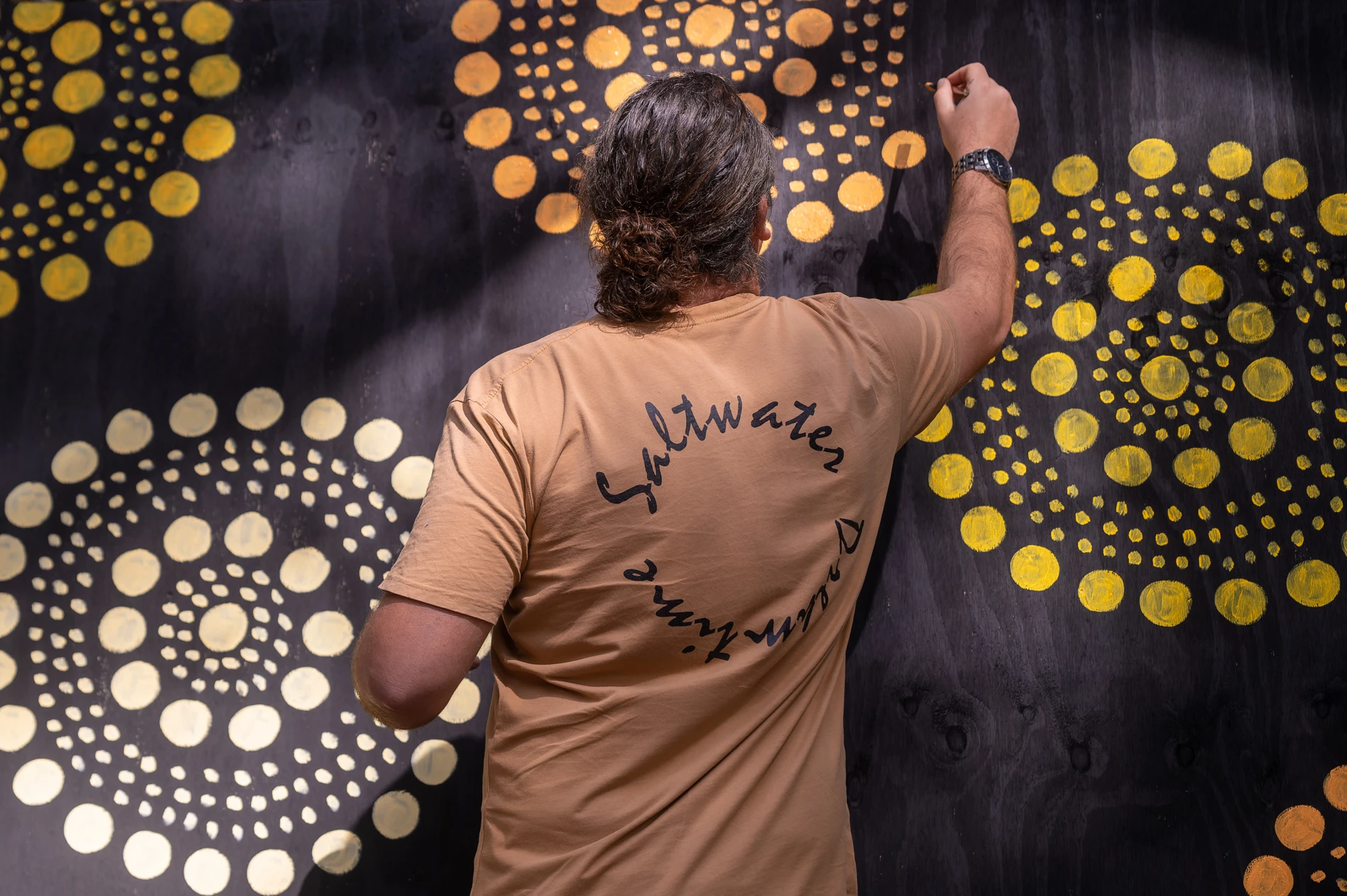 Person Painting Yellow Concentric Circles On Dark Surface, Wearing A Beige Shirt With Circular Text.