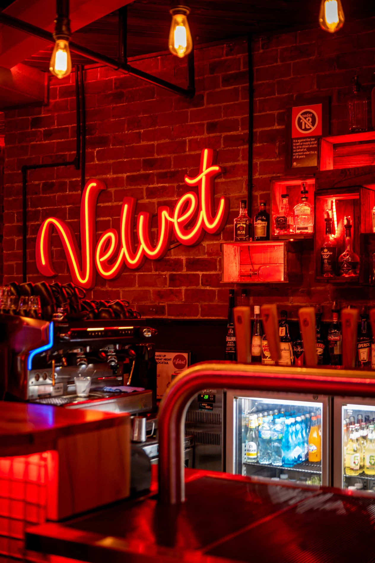 Bar Interior With Neon Velvet Sign, Liquor Shelves, Coffee Machine, And Drink Display Fridge.