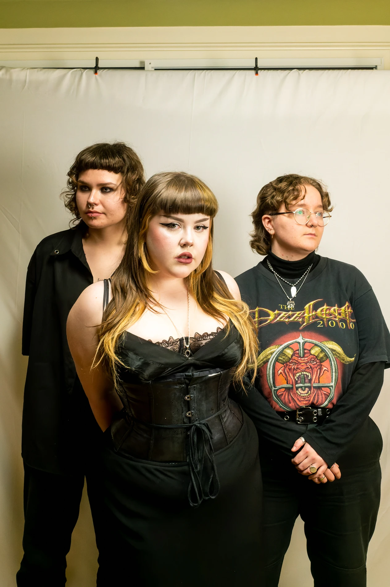 Three People In Black Attire In Front Of A Light Backdrop; Unique Hairstyles And Piercings.