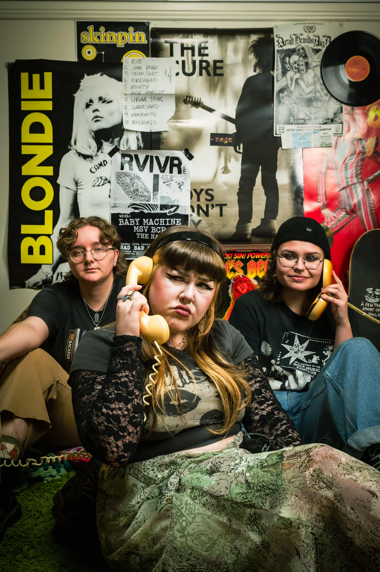 Three People Seated Indoors With Vintage Telephones, Music Posters, And A Vinyl Record On The Wall.