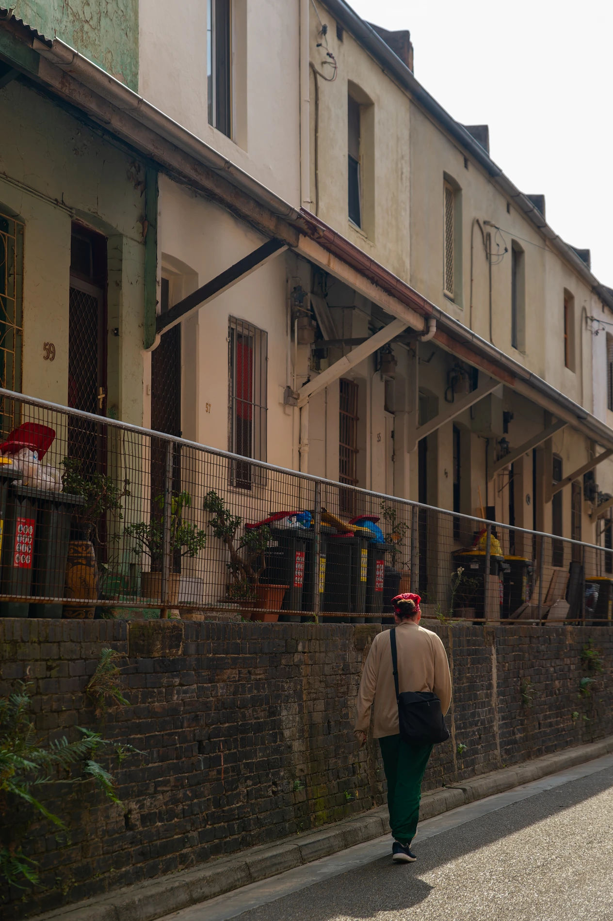 Person Walks By Weathered Terrace Houses With Bins, Plants, Fencing, And Overhead Wires Visible.