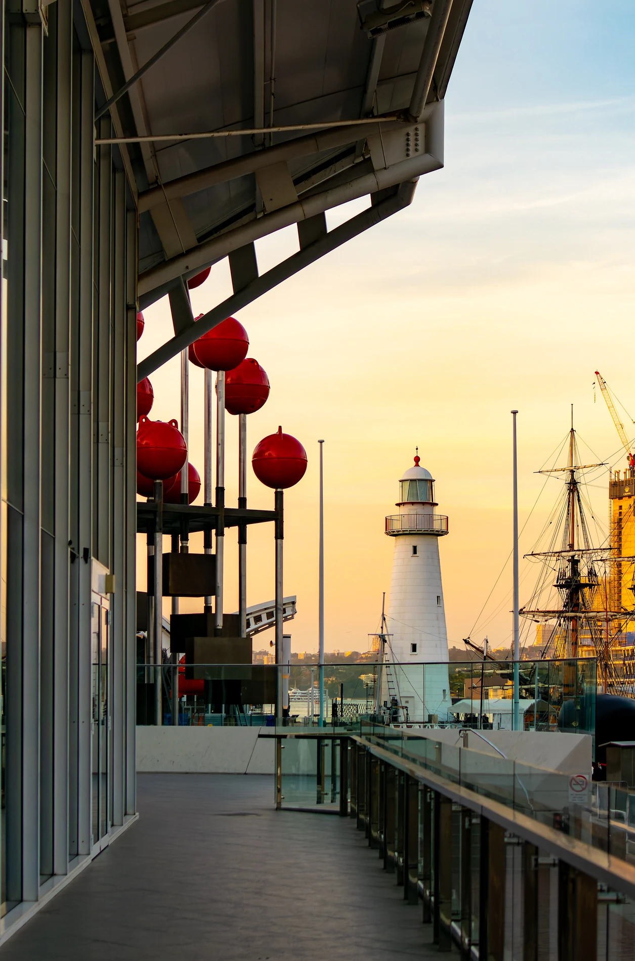 Modern Walkway With Glass Railings, Red Spheres, Lighthouse, And Historic Ship At Sunset.