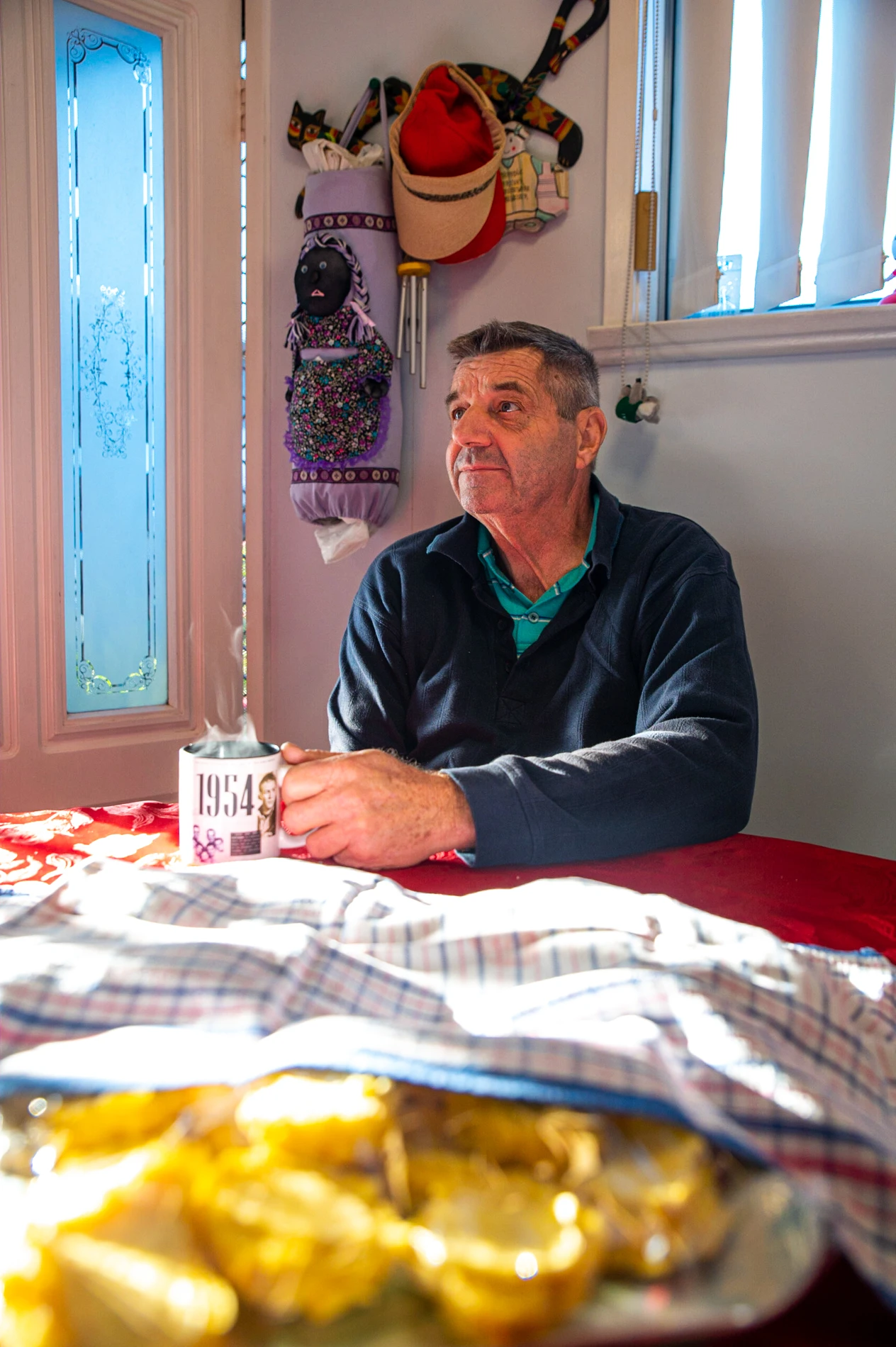 Man Holding Steaming Mug At A Table Indoors, Hats And Decor On Wall, Warm Room Lighting.