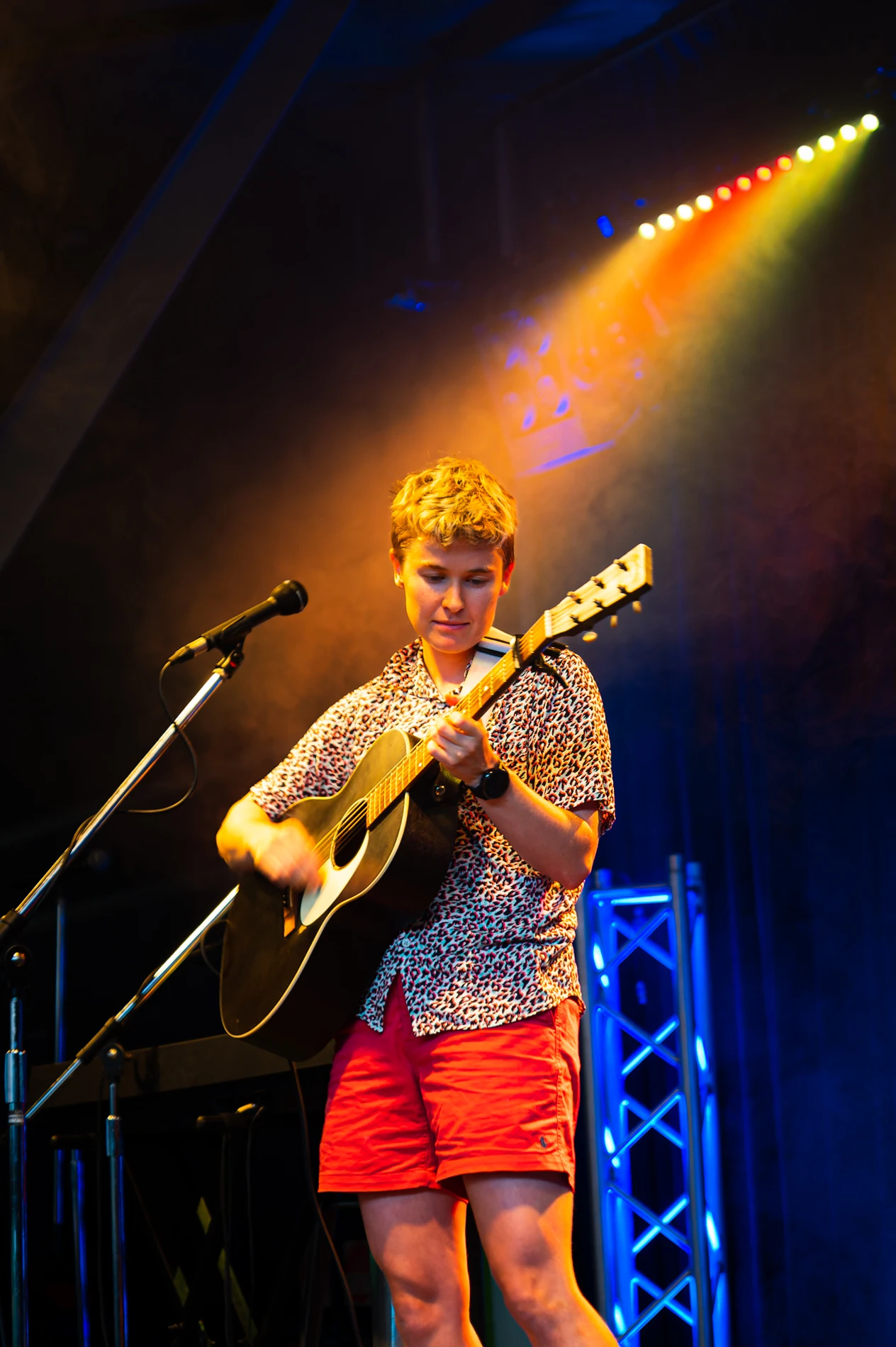 Musician On Stage With Acoustic Guitar, Patterned Shirt, Red Shorts, Colorful Lighting.