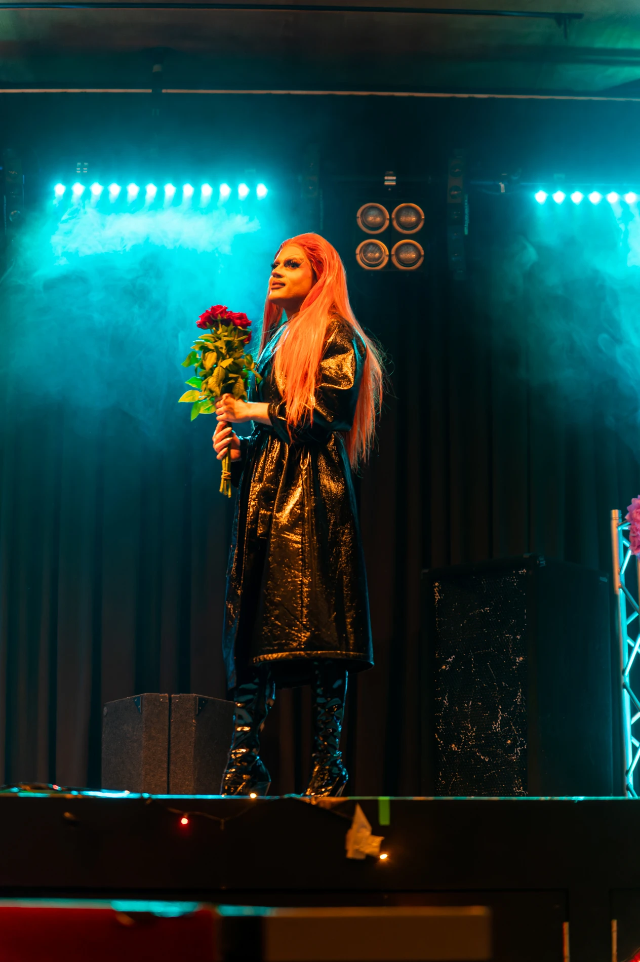 Person With Pink Hair On Stage In Blue Light, Wearing Black Coat, Holding Red Roses.