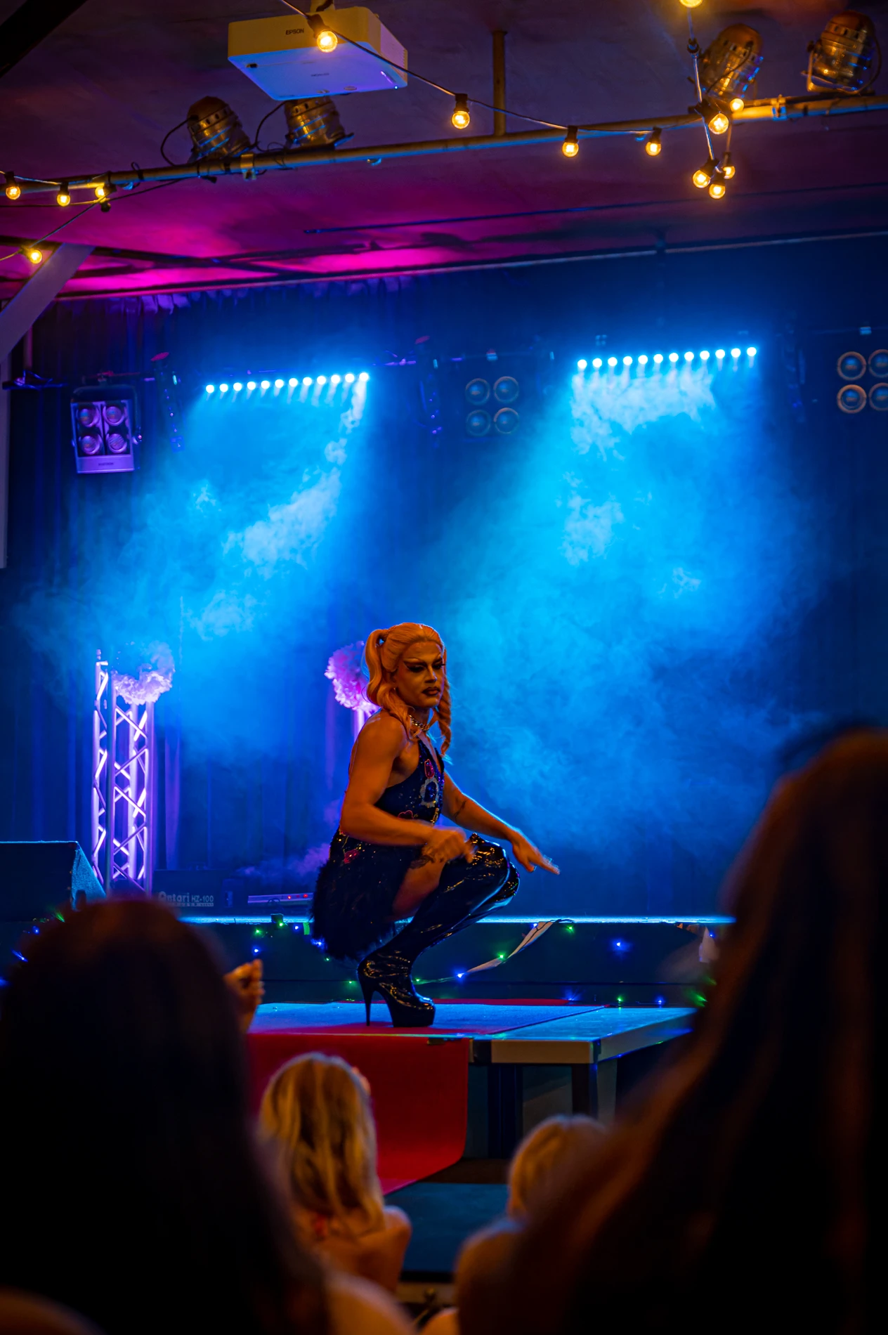 Performer Kneeling On Stage With Blue And Pink Lights, Audience Visible In Foreground.
