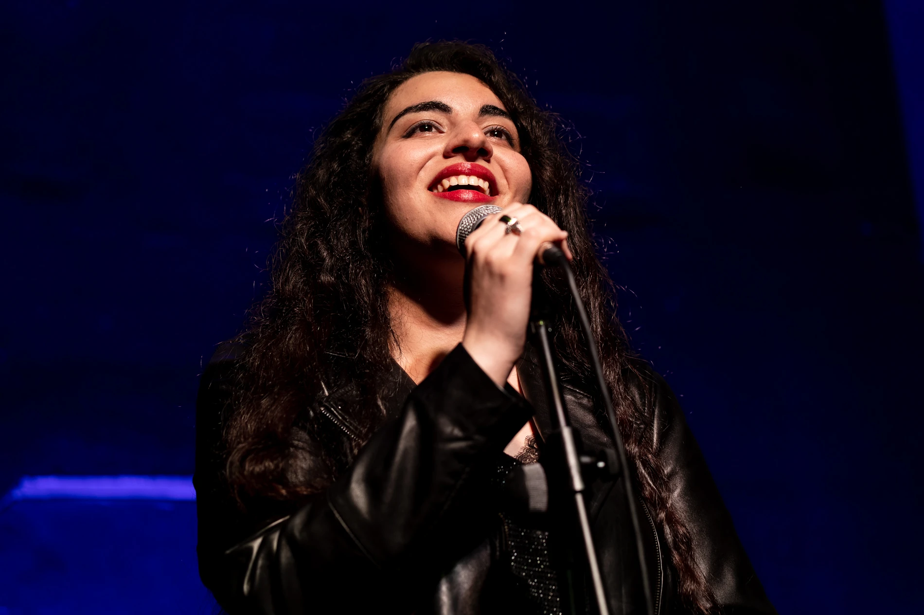 Person With Curly Hair In A Black Jacket Singing Into A Microphone On Stage.