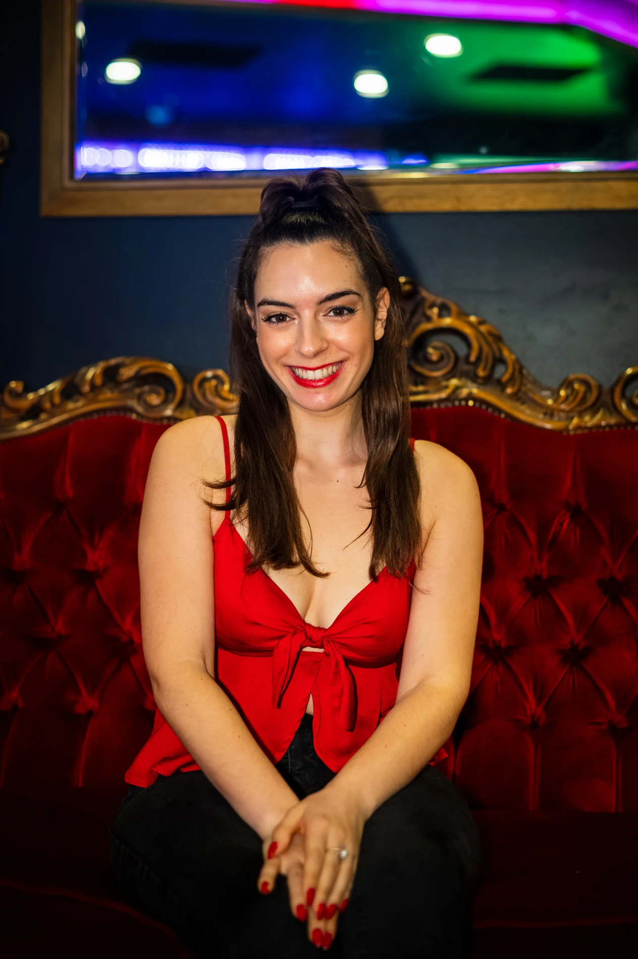 Woman With High Ponytail In Red Top, Sitting On Ornate Velvet Couch, Colorful Lights Behind.