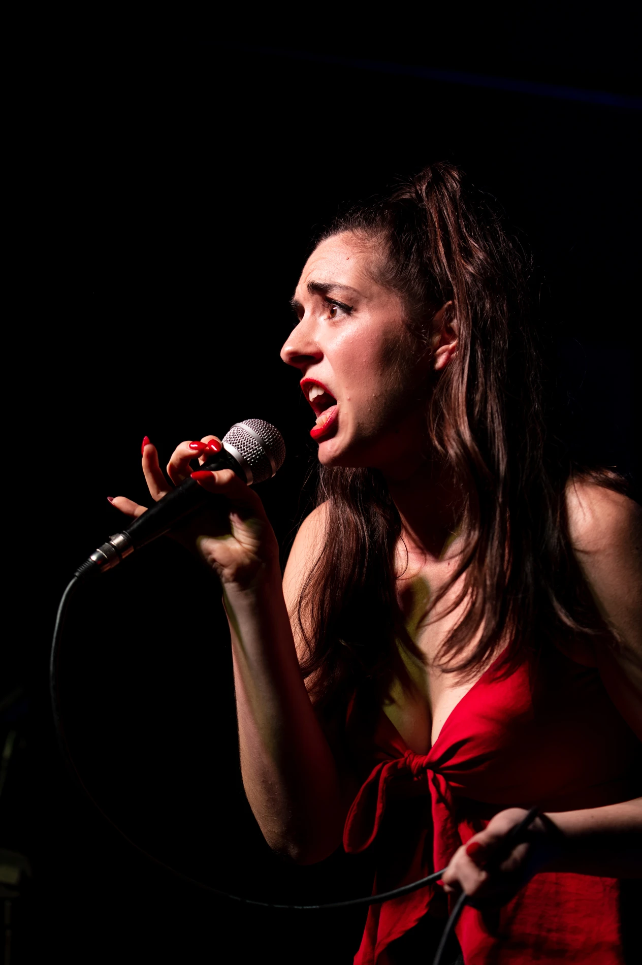 Woman In Sleeveless Red Top Passionately Singing On Stage, Holding A Microphone.
