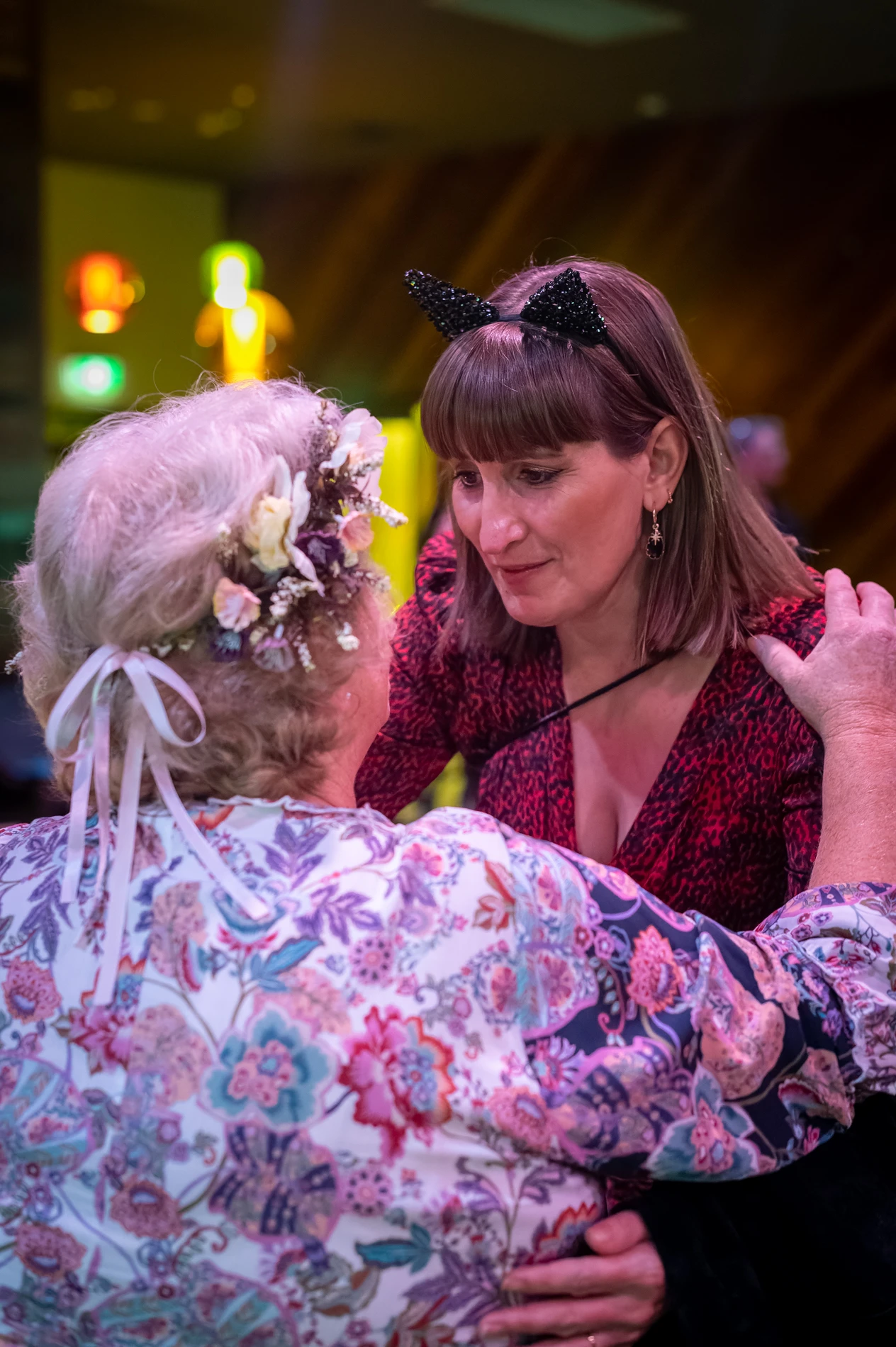 Two Women Embracing; Floral Blouse, Flower Headband, Red Dress, Cat Ears, Colourful Lights.