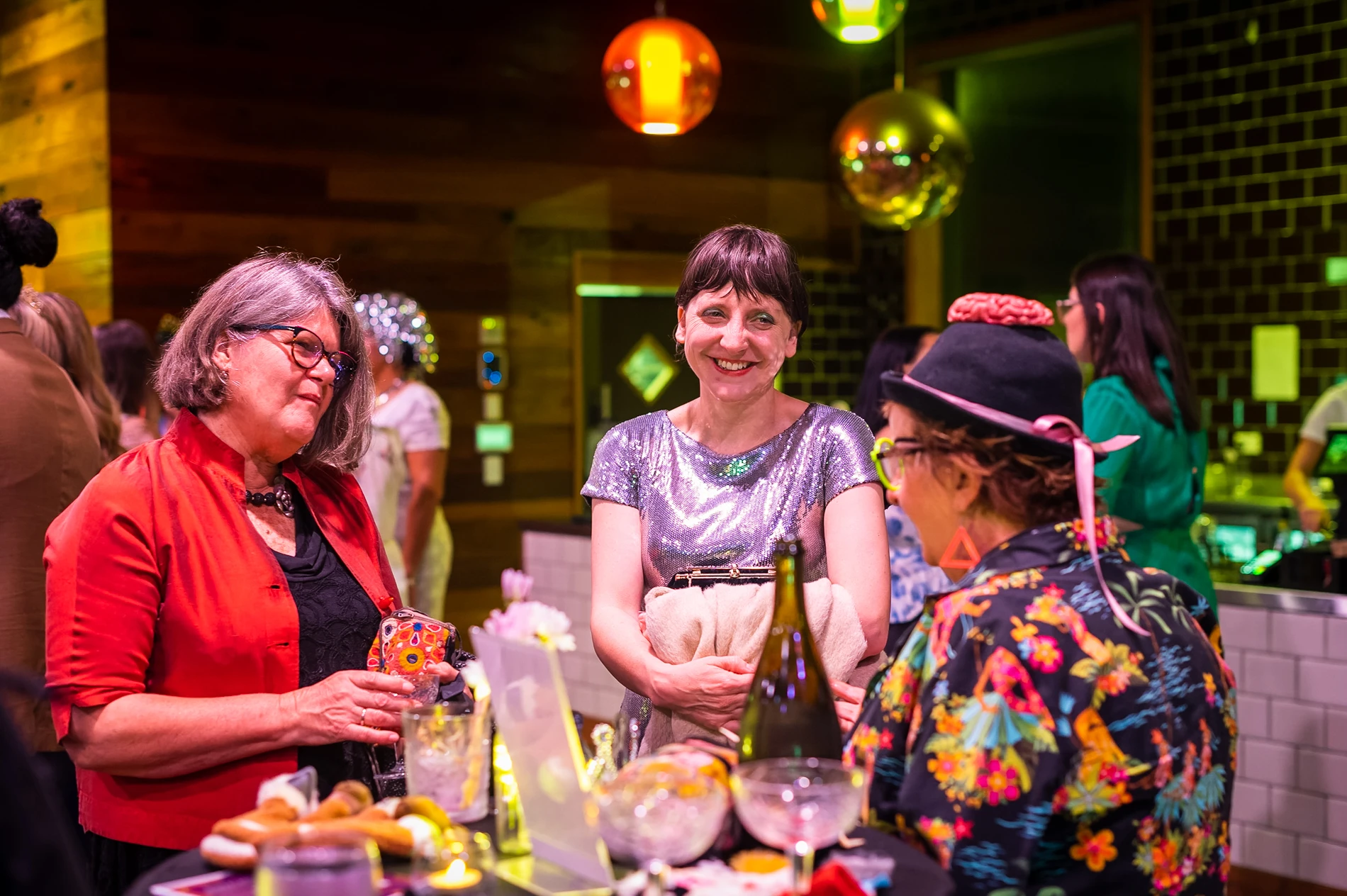 Group Socialising Indoors At A Lively Event With Drinks, Appetizers, And Colourful Decor.