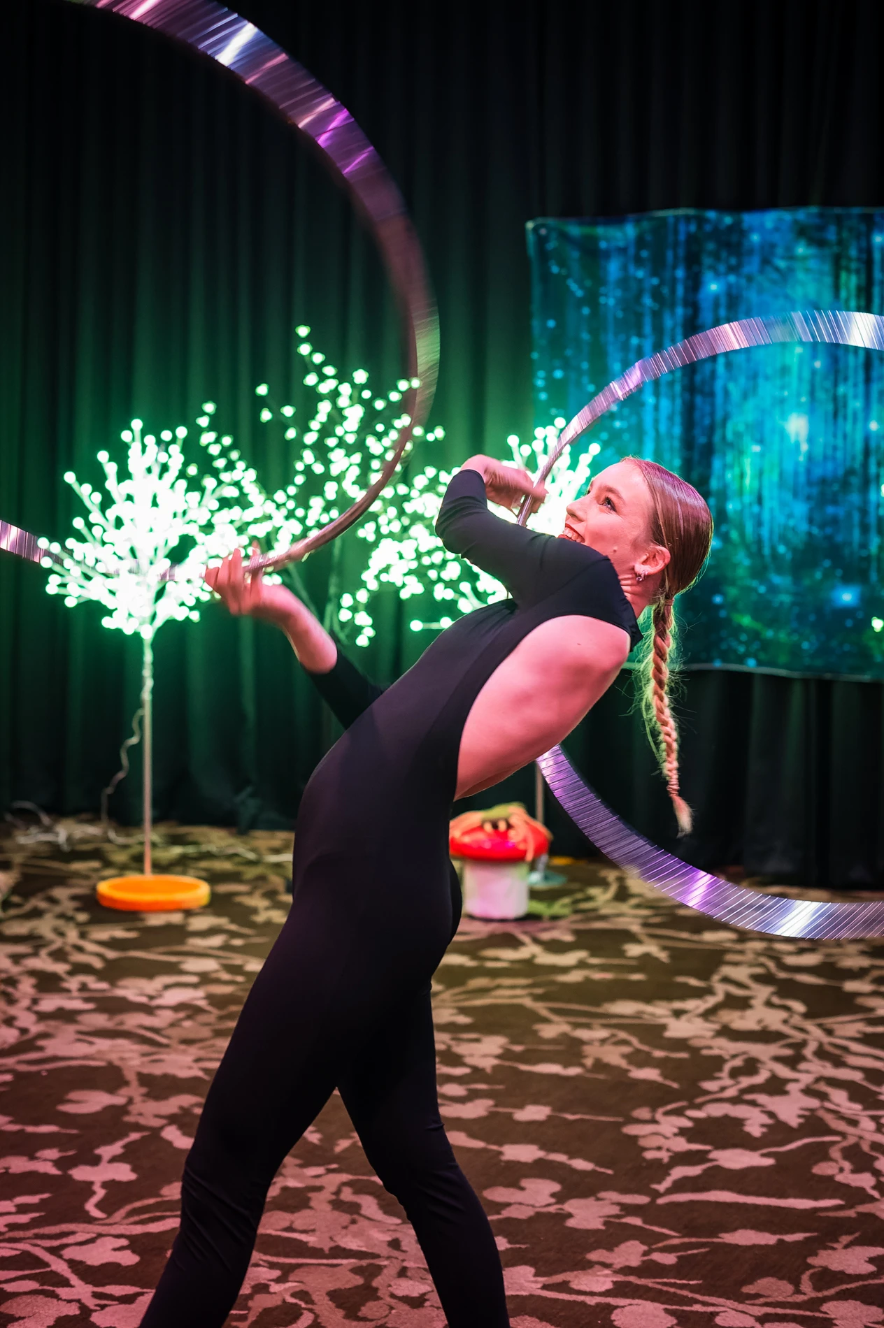 Performer In Black Bodysuit With Metallic Hoops, Dim Room, Lit Decorative Trees Behind.