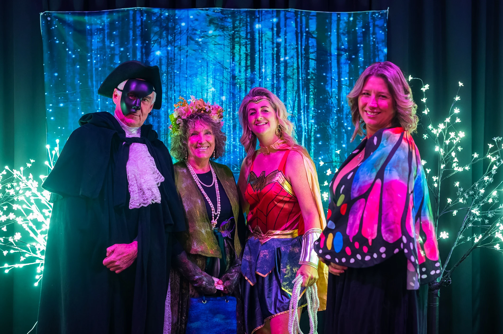 Four People In Themed Costumes Stand Before A Mystical Forest Backdrop With Colored Lights.