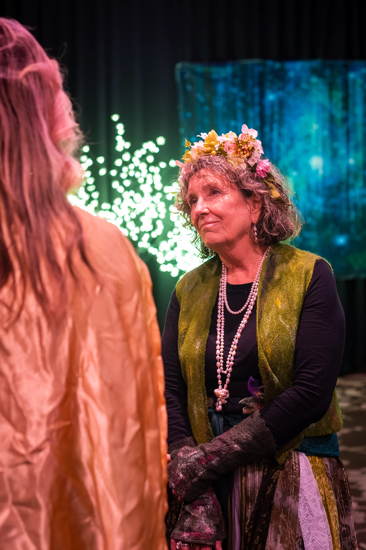 Older Woman Indoors Wearing Floral Headpiece, Layered Clothing, With Decorative Backdrop.