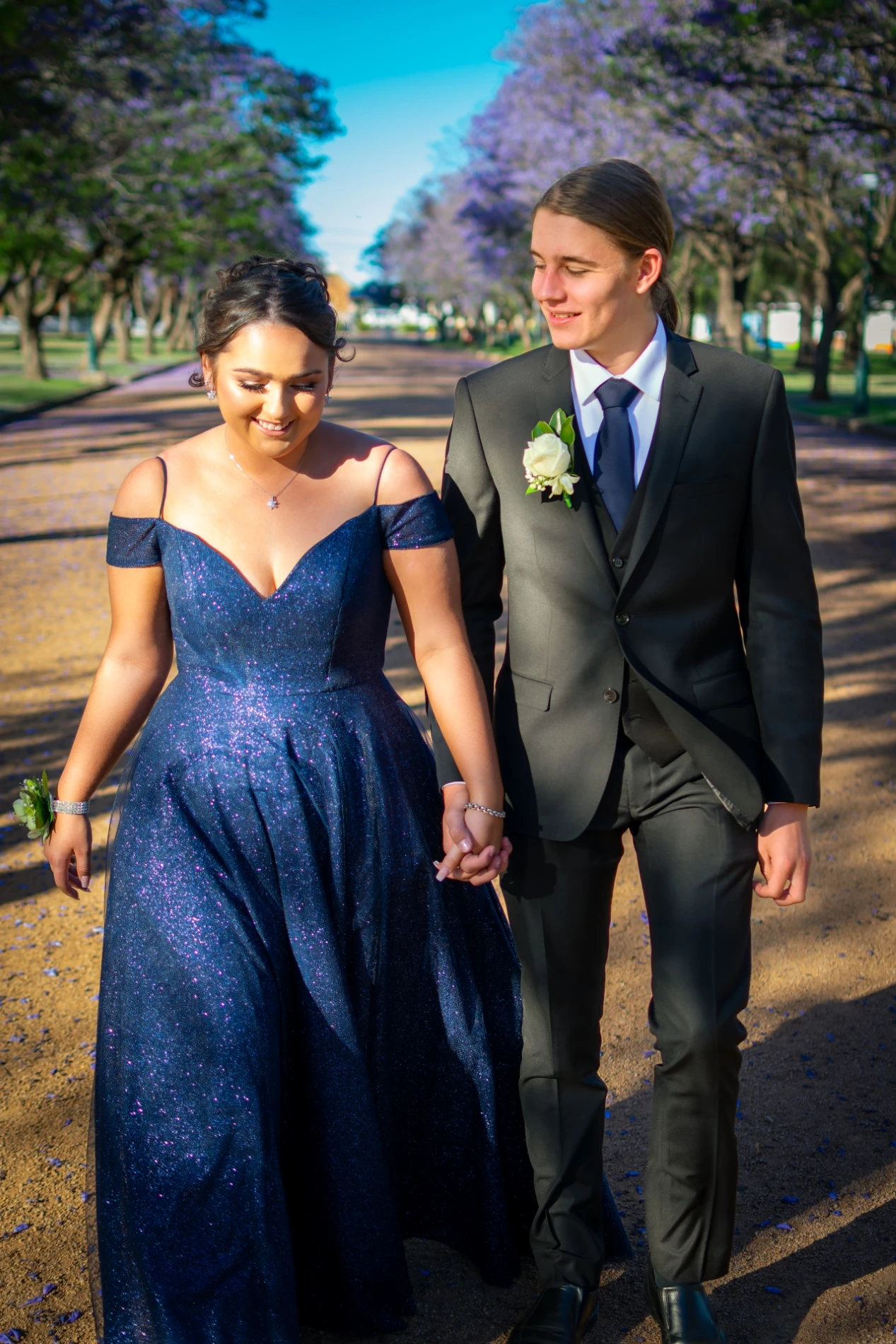 Couple In Formal Attire Walking On A Tree-Lined Path With Purple Blossoms.