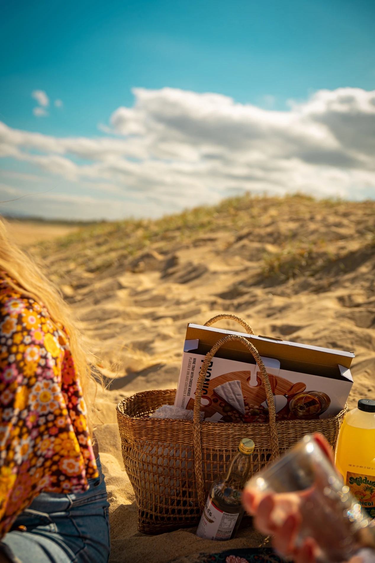 Picnic On A Sandy Beach With A Basket, Bottles, And Person In Floral Top, Clear Sky Above.