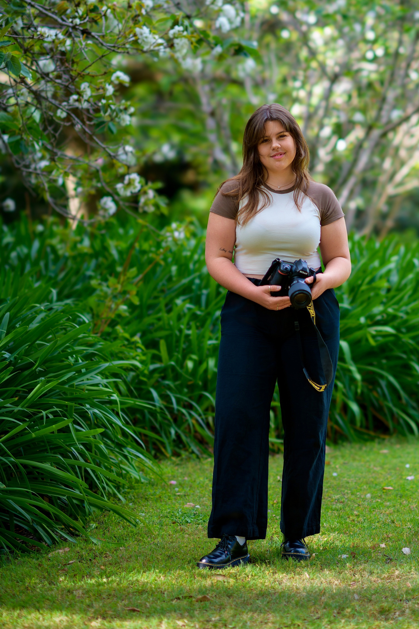 Welcome To: Neesh Photography 2.0! 7 Person In A Garden Holding A Camera, Wearing Short Sleeves And Dark Pants, With Foliage Behind.
