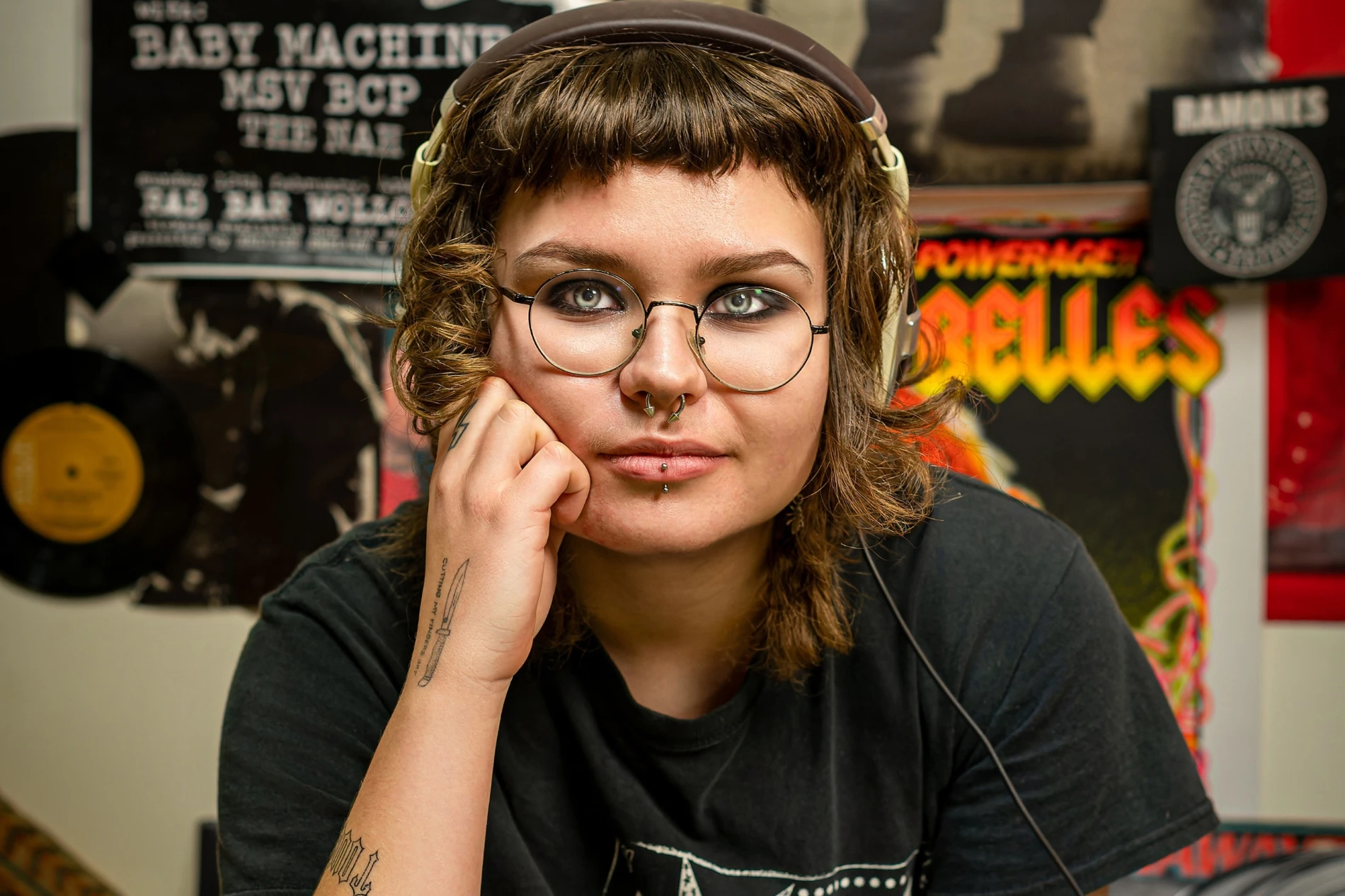 Person with glasses, headphones, short hair, piercings, in front of music posters and records.