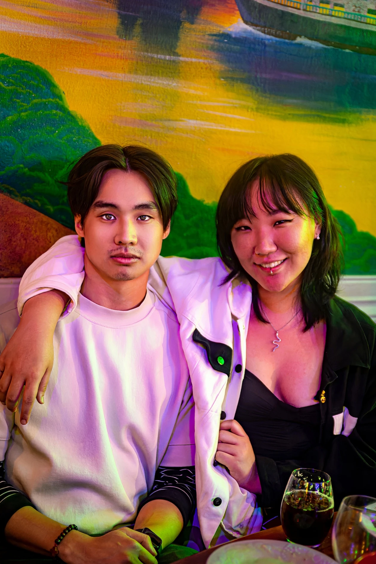 Young Couple At A Restaurant Table With Colorful Wall Art, Smiling And Relaxed With Drinks.