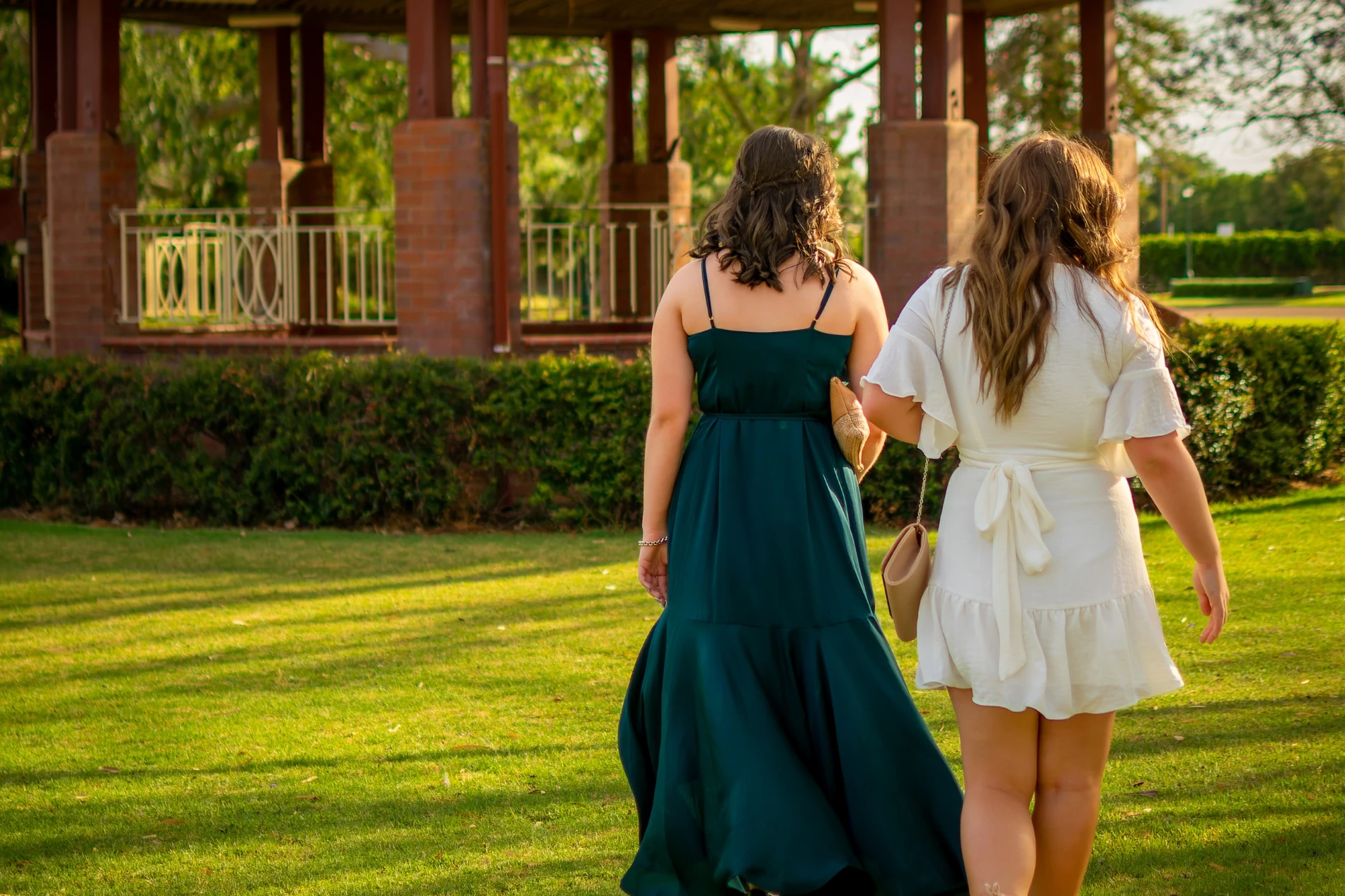 Two individuals in dresses walking on grass toward a brick gazebo surrounded by greenery.
