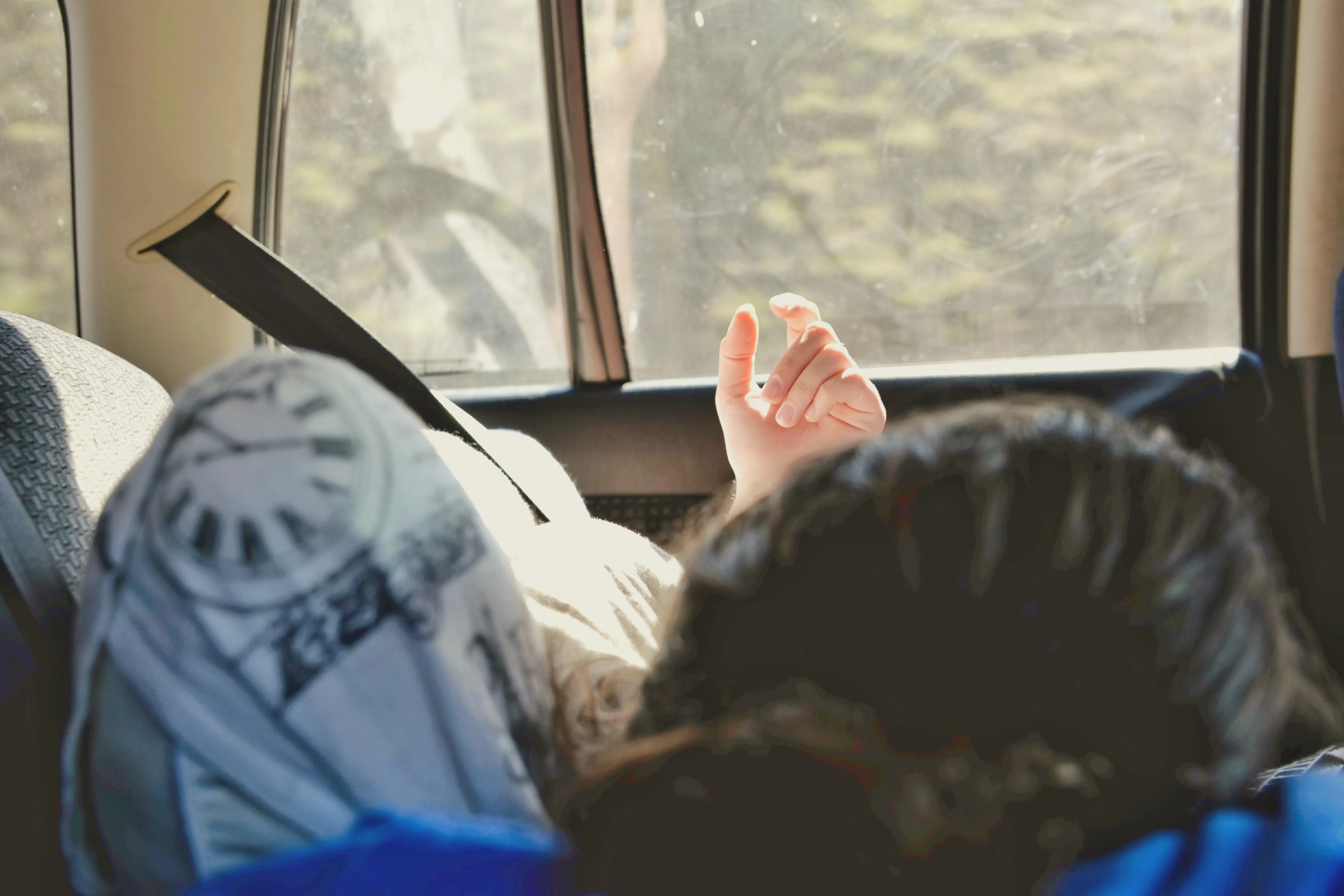 Person Reclining In Car, Hand Raised, Sunlight Filtering In, Clock-Patterned Cushion Visible.