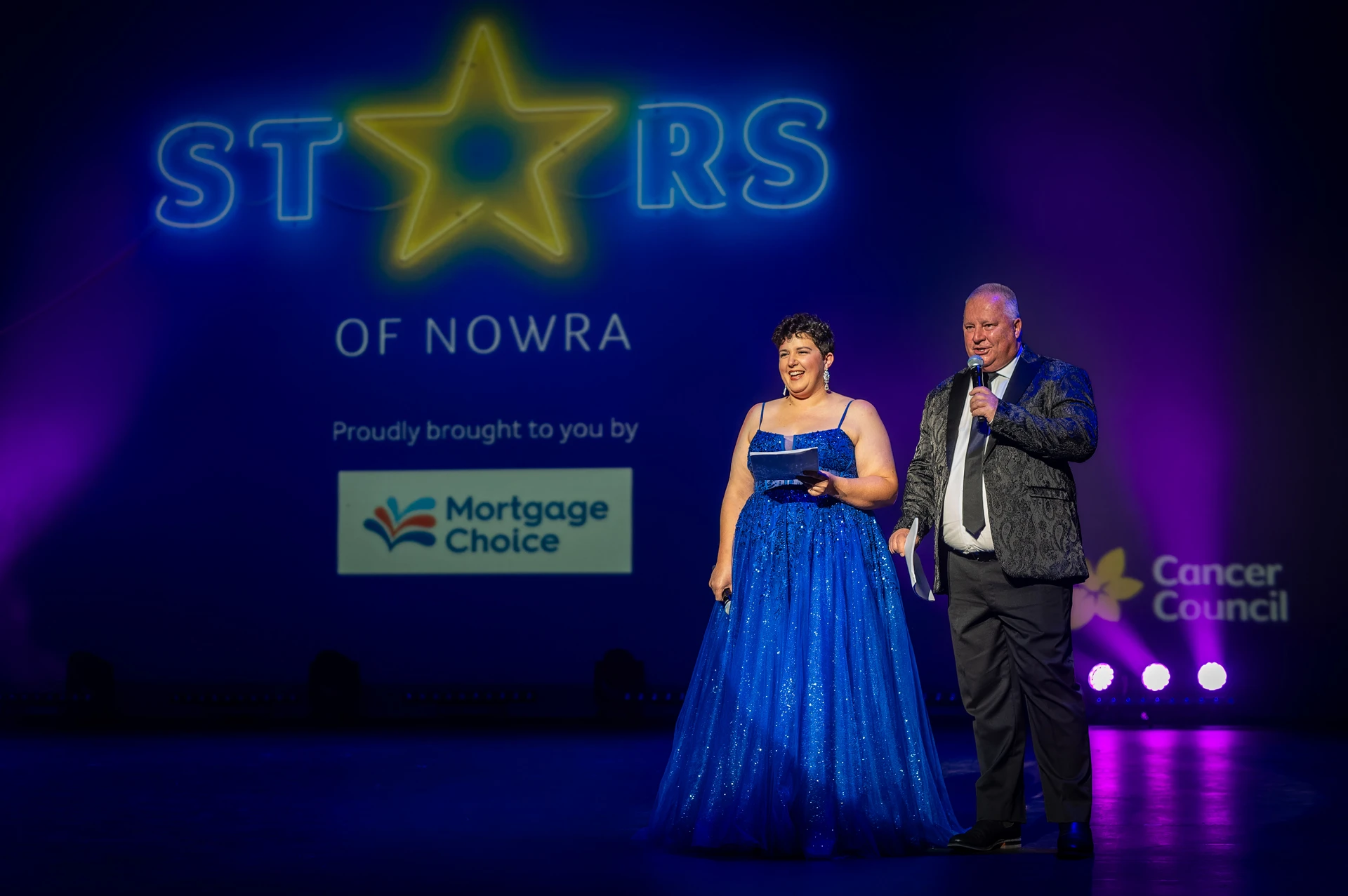 Two people on stage at "Stars of Nowra," wearing formal attire, with sponsor logos behind.