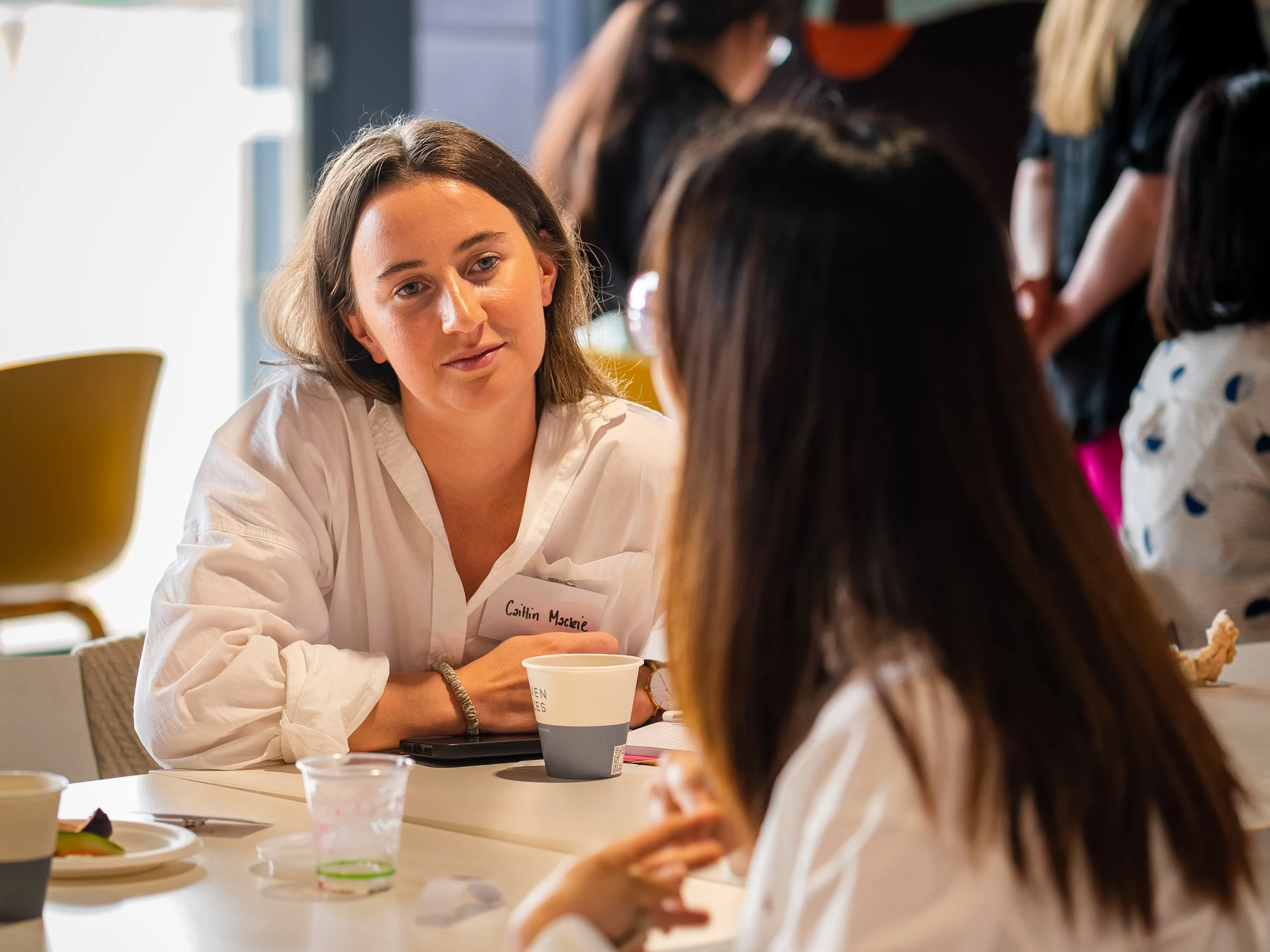 Two women at a table talking, one with a white shirt and name tag, others in the background.