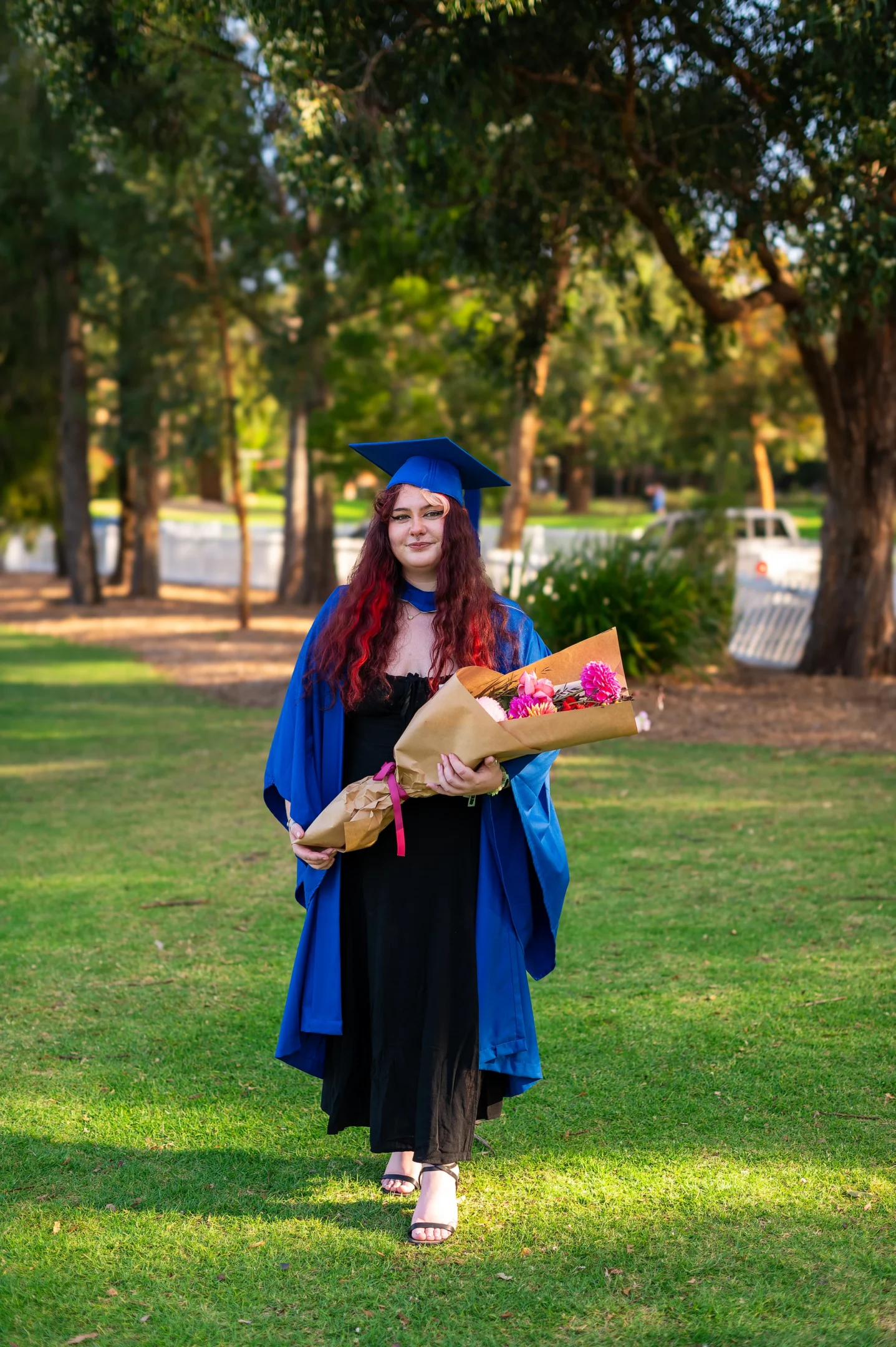 Graduate In Blue Cap And Gown Holding Bouquet On Grass With Trees In The Background.