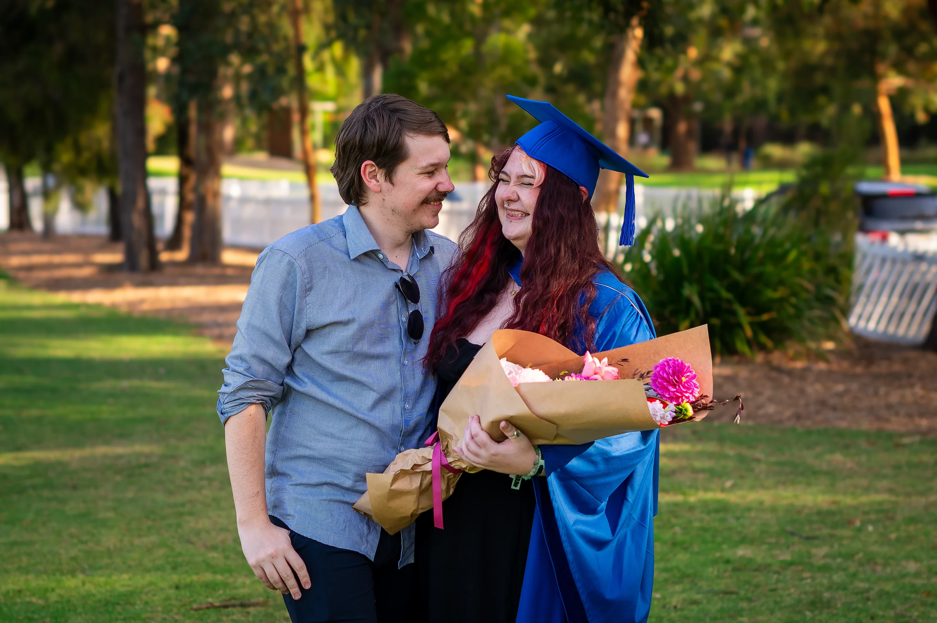 Graduate In Cap And Gown Holds Flowers, Standing With A Casually Dressed Individual Outdoors.