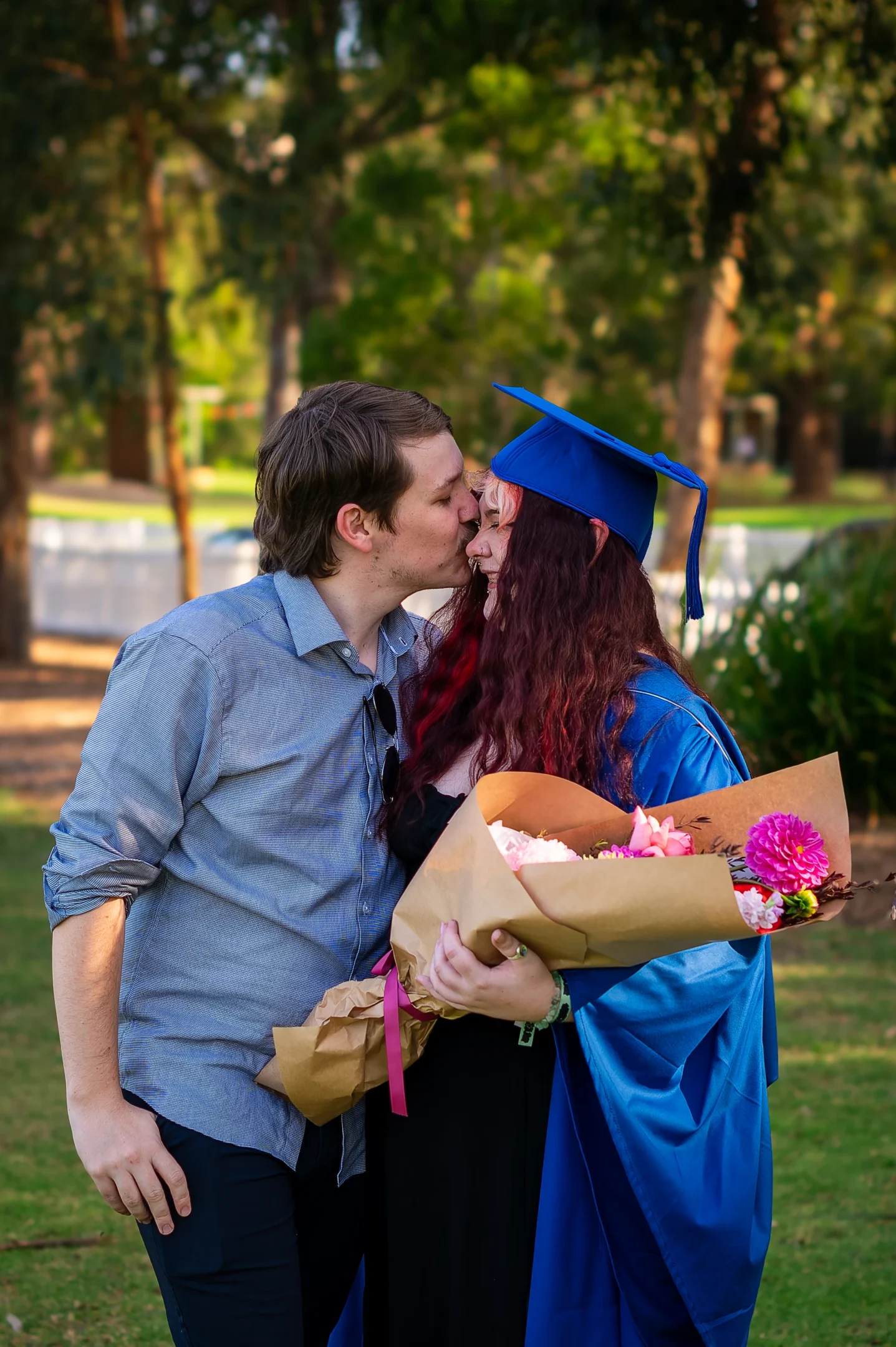 Graduate In Blue Cap And Gown Holds Flowers, Kissed On Forehead Outdoors.