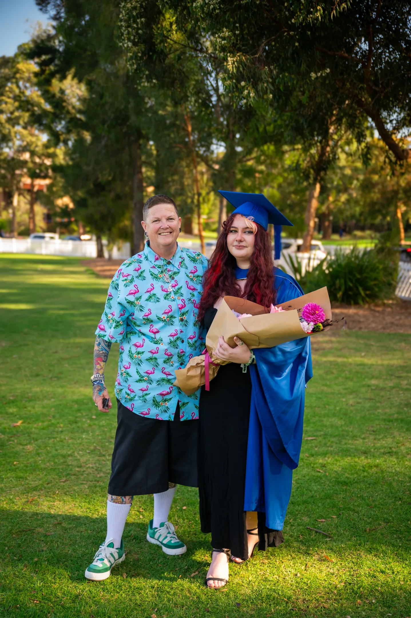 Two People Outdoors; One In Flamingo Shirt, Other In Blue Graduation Gown, Holding Flowers.