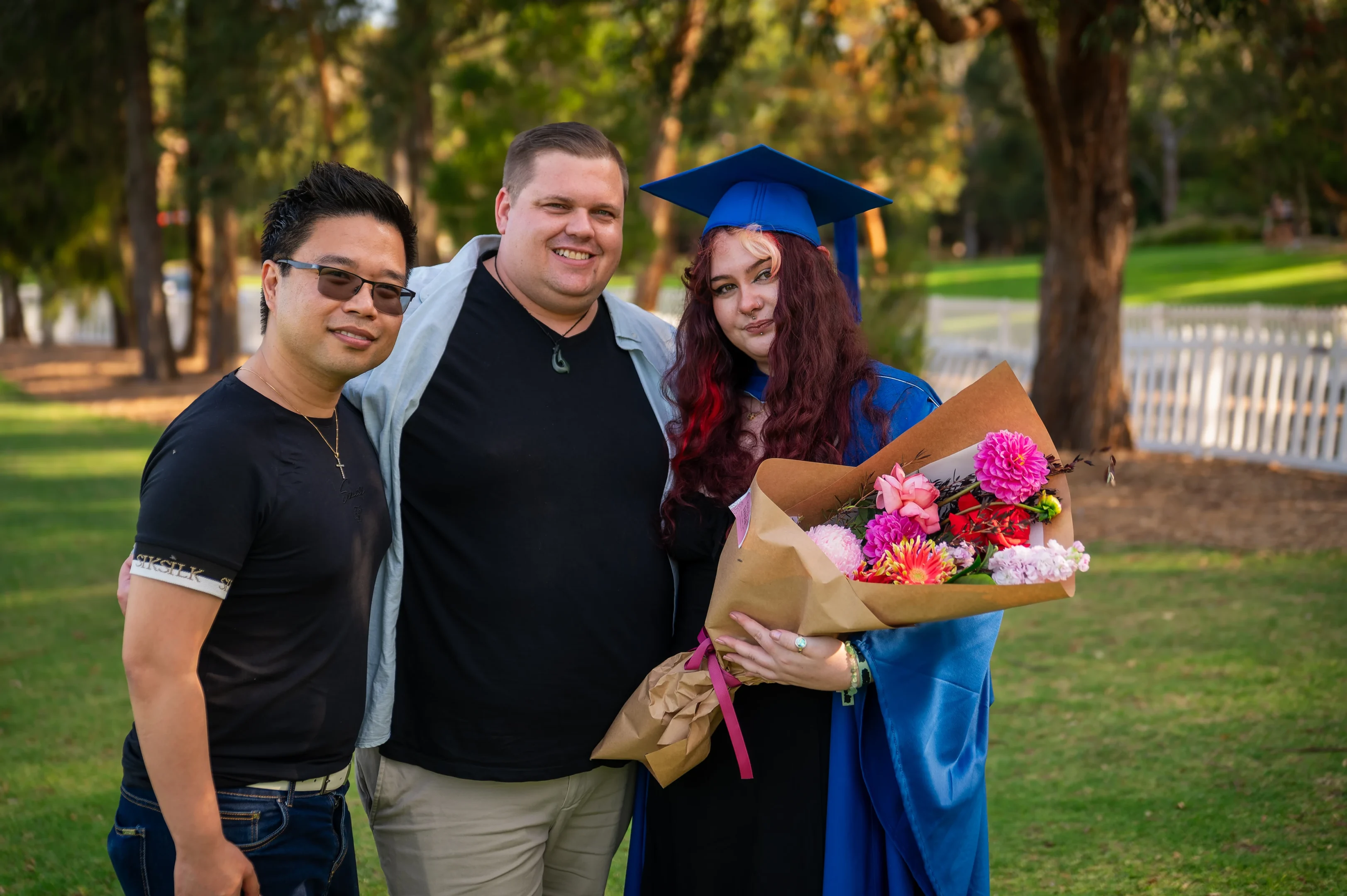 Three People Outside, One In Blue Graduation Attire Holding Flowers, Trees And A Fence Behind.